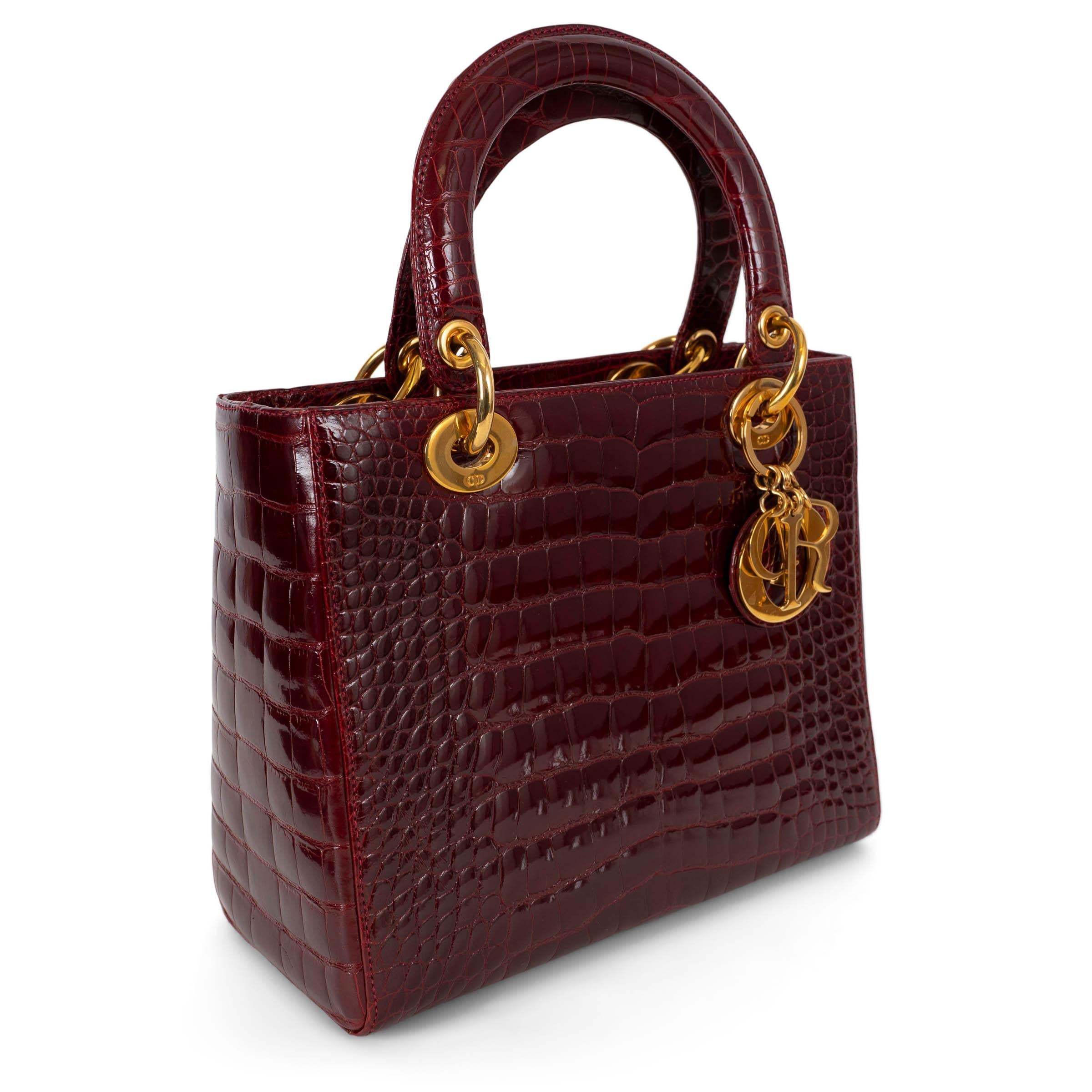 Ce sac Christian Dior Medium Lady Dior, 100% authentique, est réalisé en cuir de crocodile bordeaux brillant, d'un éclat luxueux. La silhouette structurée est agrémentée de deux poignées rigides et d'une bandoulière optionnelle pour un style