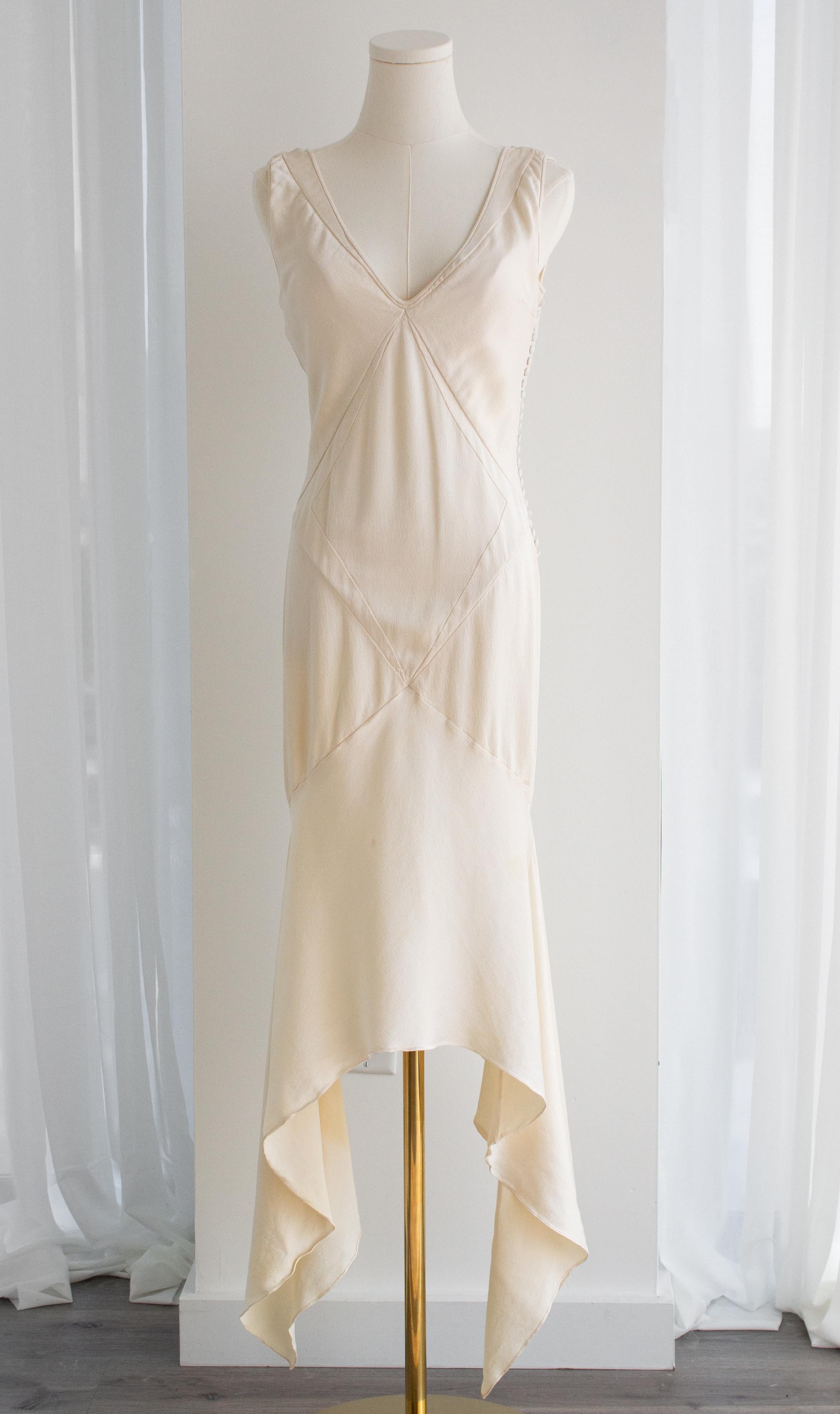 This vintage Christian Dior dress, designed by John Galliano, is crafted from luxurious textured silk in a creamy ivory-white shade. The fabric showcases a subtle water ripple effect, adding a unique, flowing texture to the bias-cut design. The bias