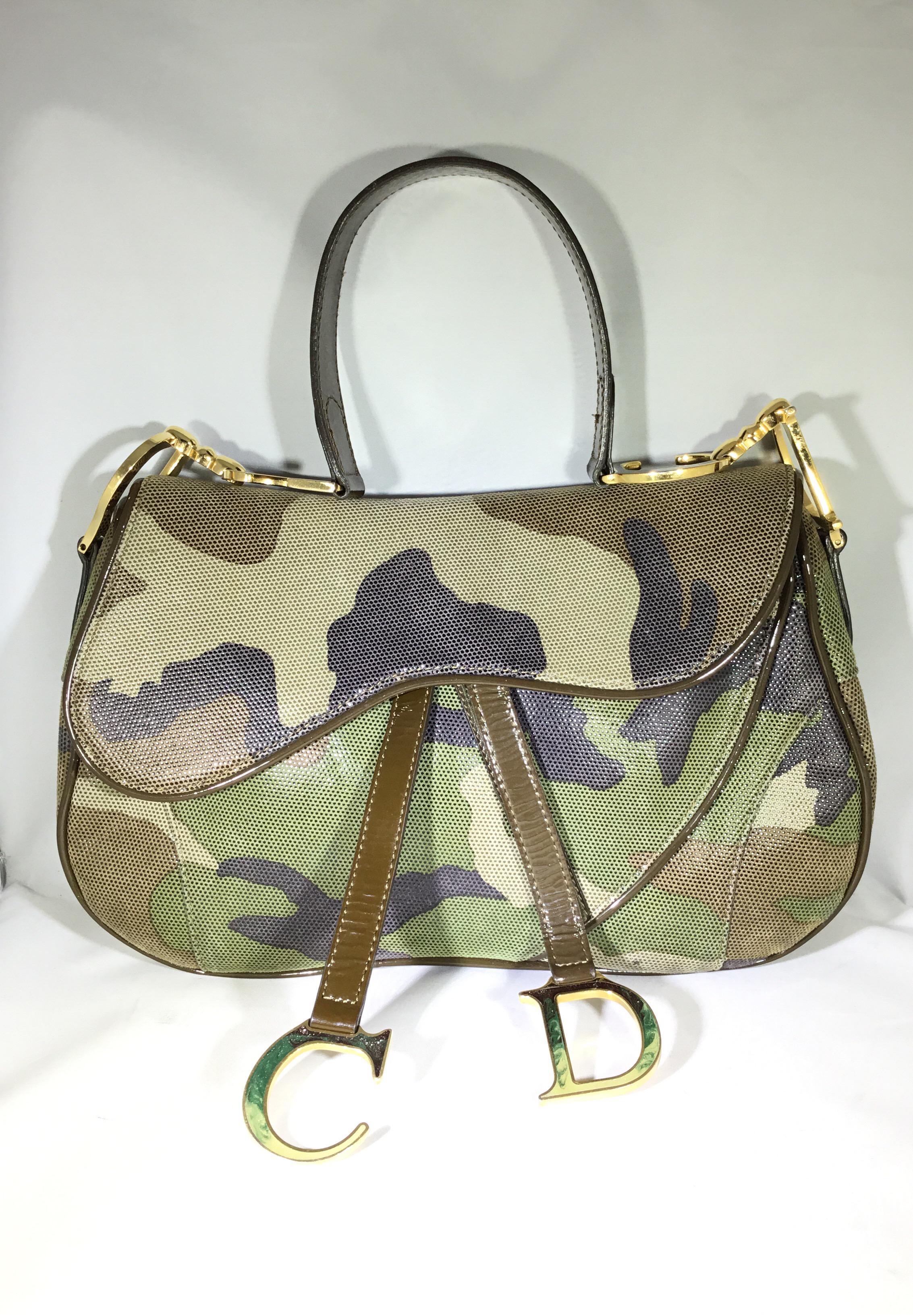 Christian Dior camouflage saddle shoulder bag has gold-tone hardware throughout, bag is made with a perforated suede atop a patent base with patent piping and shoulder strap. Velcro strap closure with a “CD” metal initial detail. Nylon lining with