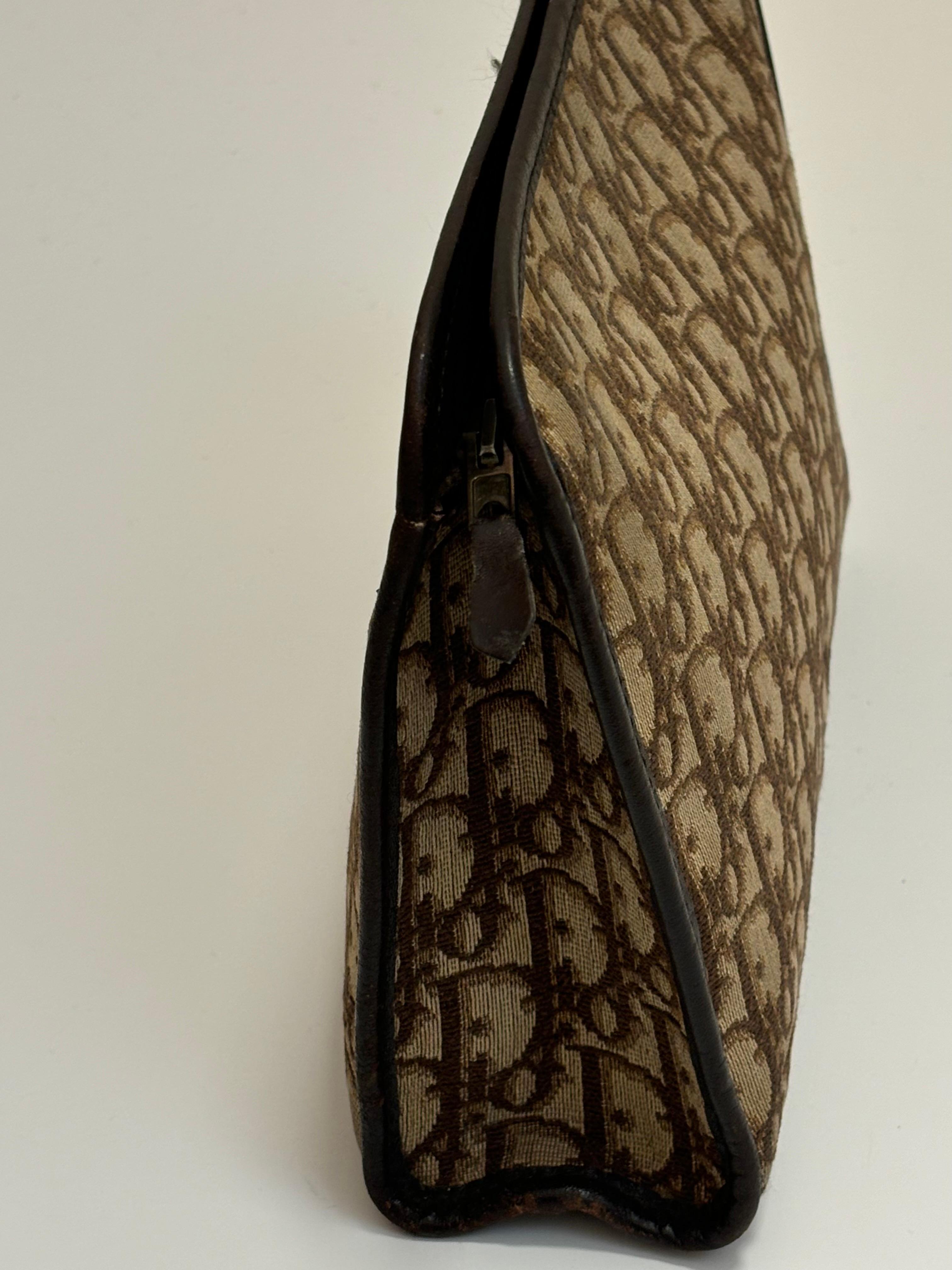 Réalisée en toile et garnie de cuir, cette pochette Christian Dior arbore le célèbre motif CD trotter. Les couleurs sont le marron et le beige. La doublure est en cuir beige et il y a deux grands compartiments à fentes. 
La pochette est dotée d'une
