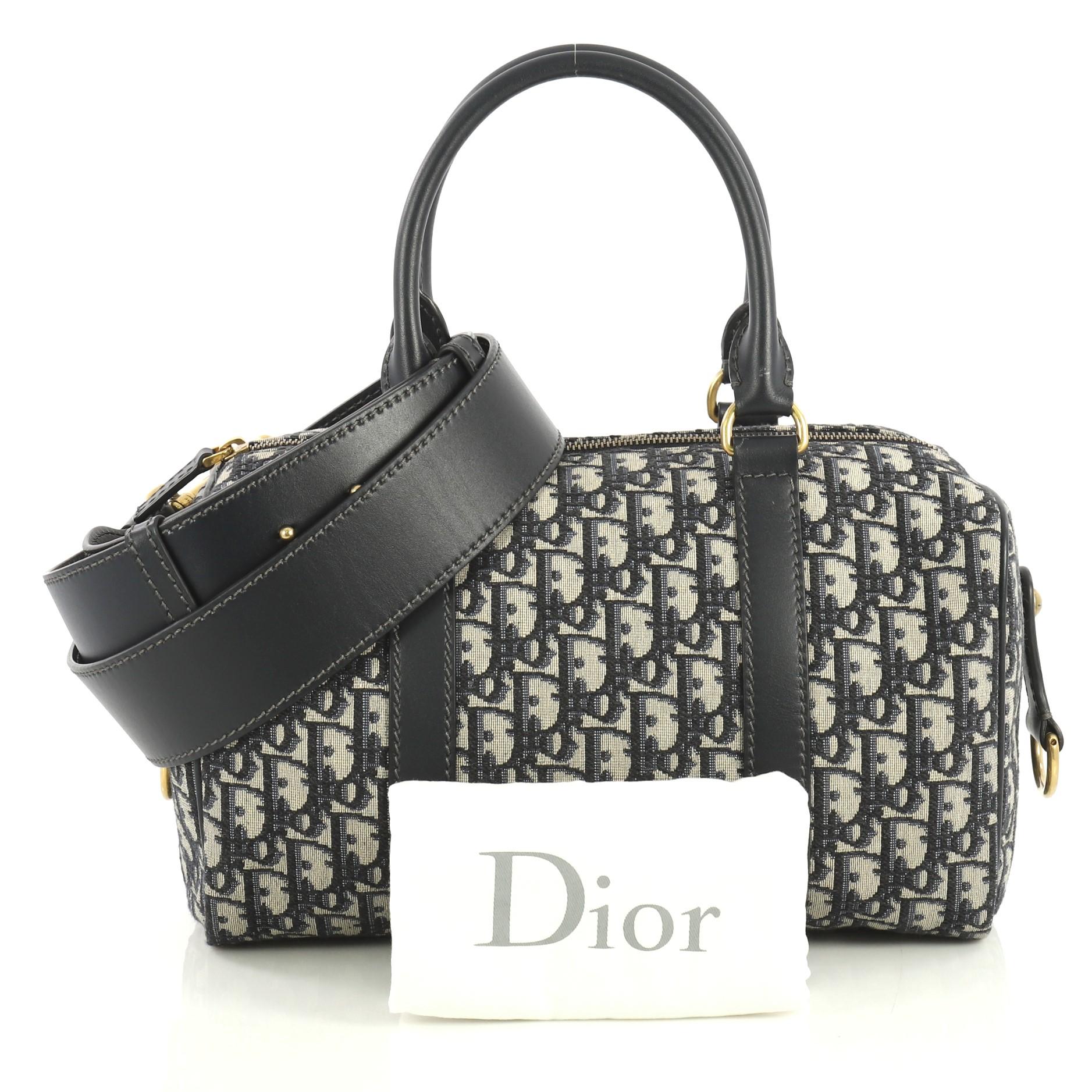 This Christian Dior Convertible Boston Bag Oblique Canvas Medium, crafted from blue and neutral canvas, features dual rolled handles, leather trim, and aged gold-tone hardware. Its zip closure opens to a neutral fabric interior with zip pocket.