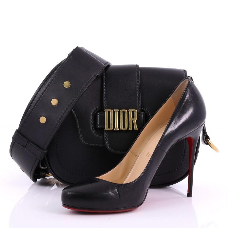 This Christian Dior D-Fence Saddle Bag Leather Small, crafted from black leather, features removable and adjustable shoulder strap, gold-tone DIOR buckle fastening, and gold-tone hardware. Its tuck flap closure opens to a black suede interior with