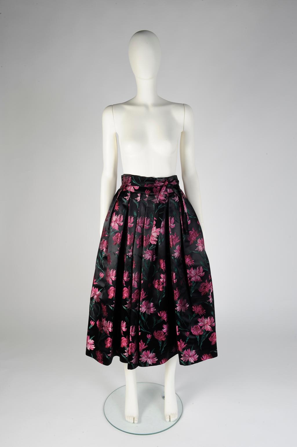 Dieser seltene Midirock von Christian Dior aus den frühen 50er Jahren ist leicht ausgestellt und verfügt über einen hochgezogenen Bund und einen tonalen Schärpengürtel. Dieser Rock aus glänzendem Seidendamast zeigt raffinierte rosa Nelken auf