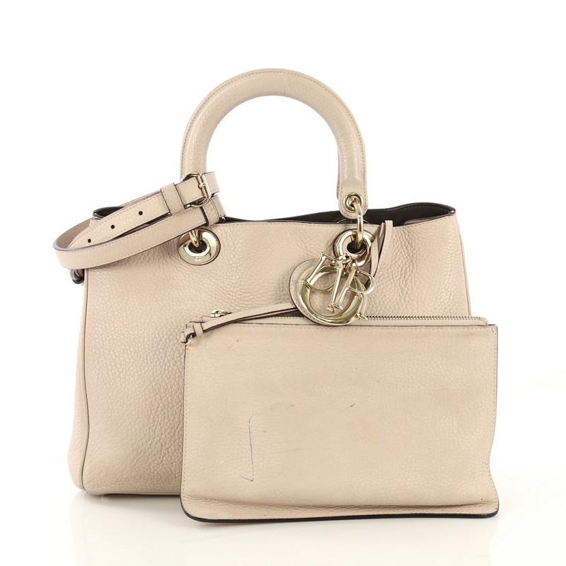 This Christian Dior Diorissimo Tote Pebbled Leather Medium, crafted from pink pebbled leather, features short dual handles with Dior charms, side snap buttons, protective base studs, and gold-tone hardware. Its magnetic snap button closure opens to