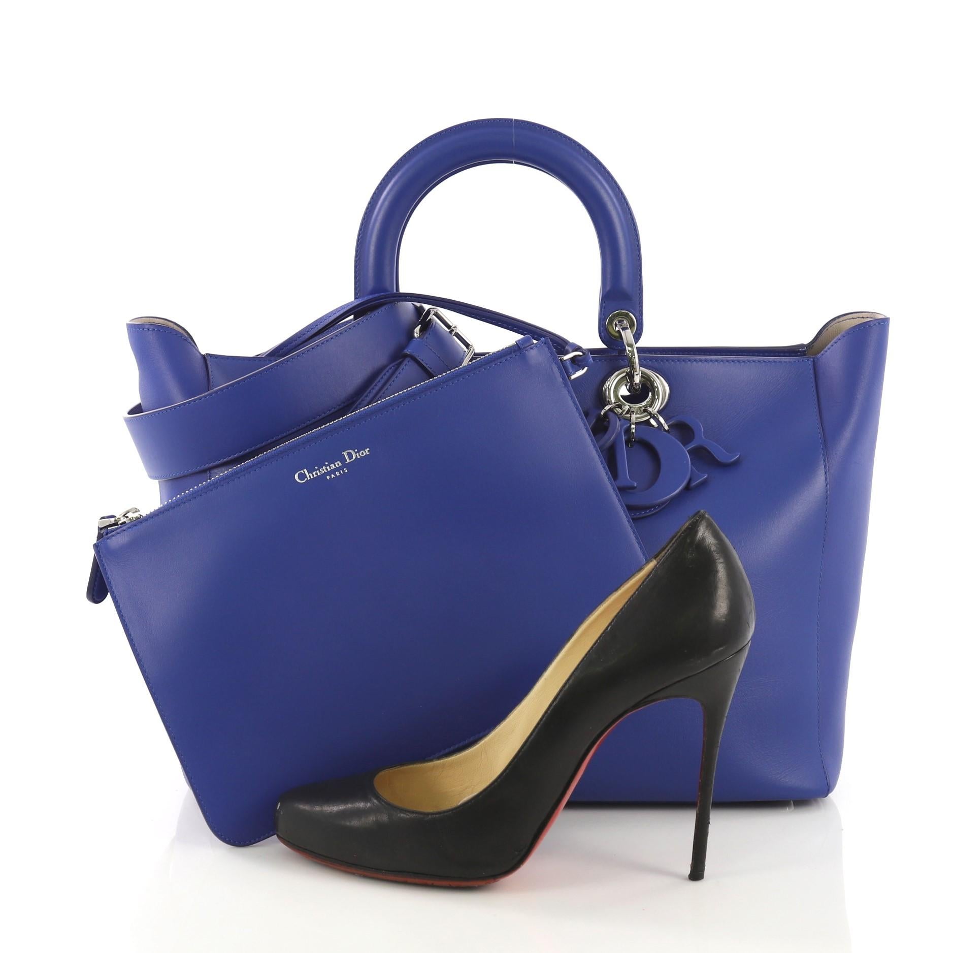 This Christian Dior Diorissimo Zip Tote Smooth Calfskin Large, crafted from blue smooth calfskin leather, features smooth short dual handles with Dior charms, protective base studs and silver-tone hardware. It zip closure opens to a beige leather