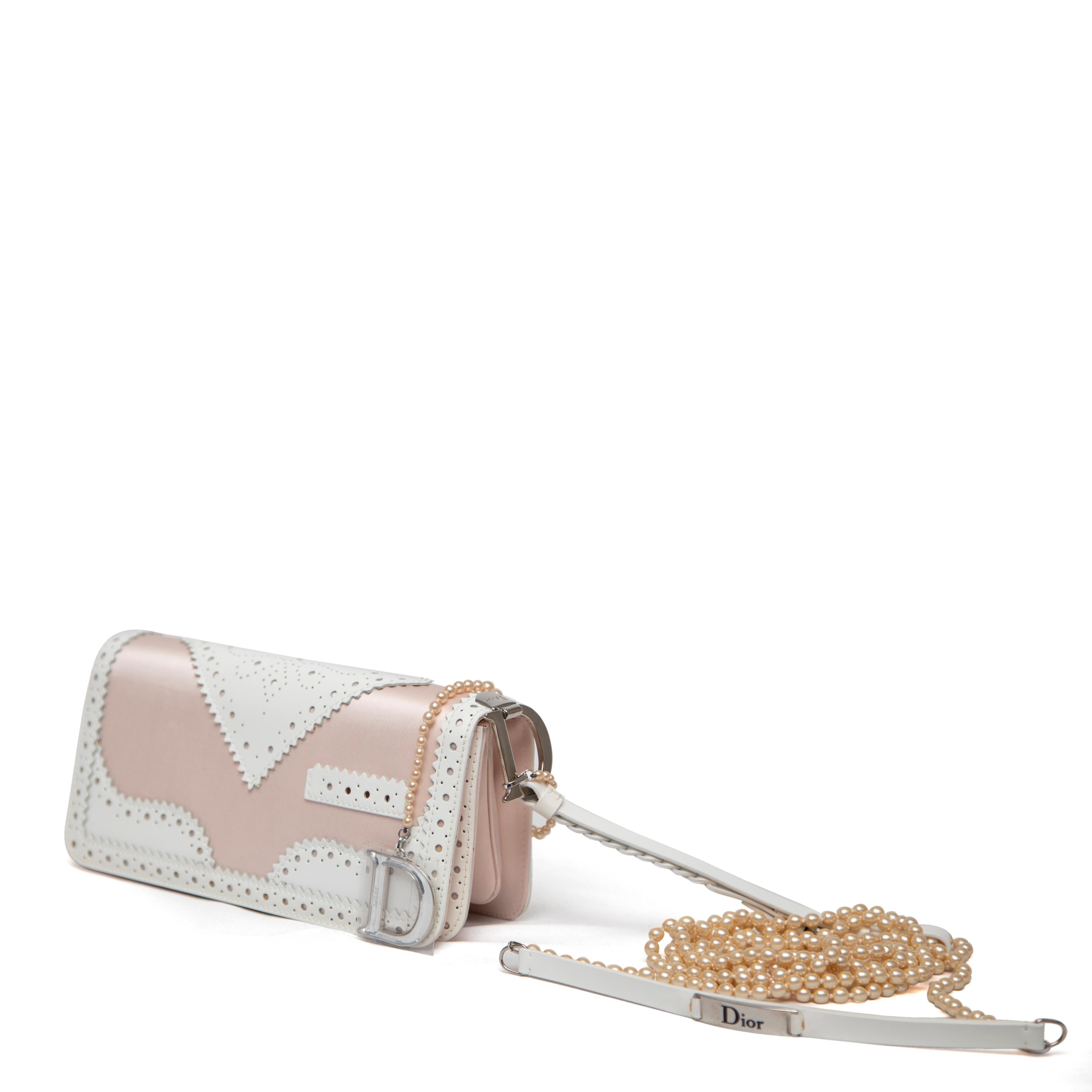 This Christian Dior D'Trick Shoulder Bag is a stunning baguette-style piece crafted from luxurious pink satin, accented with white leather trim and silver-tone hardware. It features a braided wrist strap adorned with a logo charm, as well as a pearl