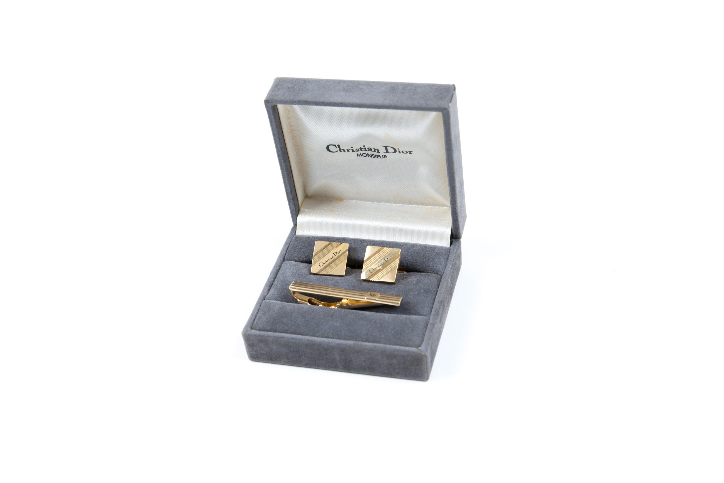 Goldfarbenes Set aus Krawattenklammer und Manschettenknöpfen von Christian Dior mit quadratischer Form, diagonalen Streifen und einem schwarzen Christian Dior-Stempel in der Mitte. Sie werden als Paar + Krawattenzubehör geliefert. 
Wird in der