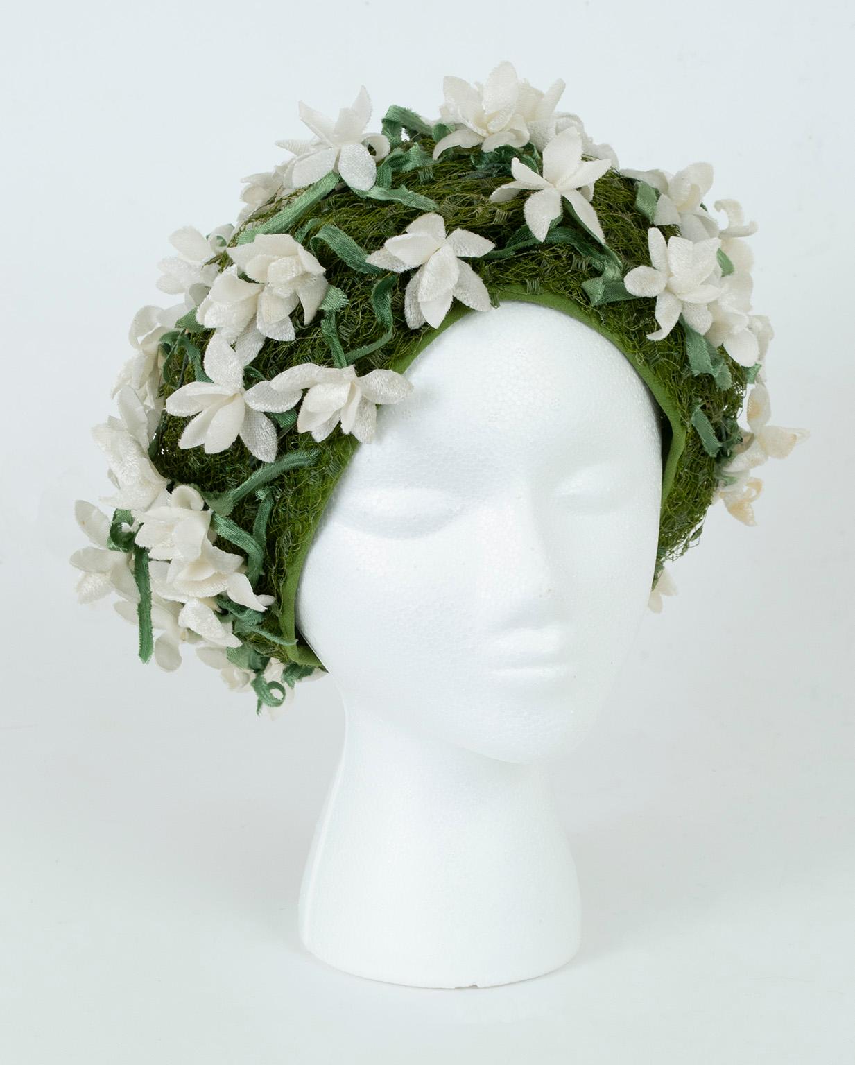 Accessoire de rêve, cet incroyable chapeau Dior utilise de la soie verte et du voilage pour simuler un tapis d'herbe, tandis que des gardénias et des feuilles de velours de soie articulés recouvrent sa surface. Sa silhouette, ses couleurs et ses