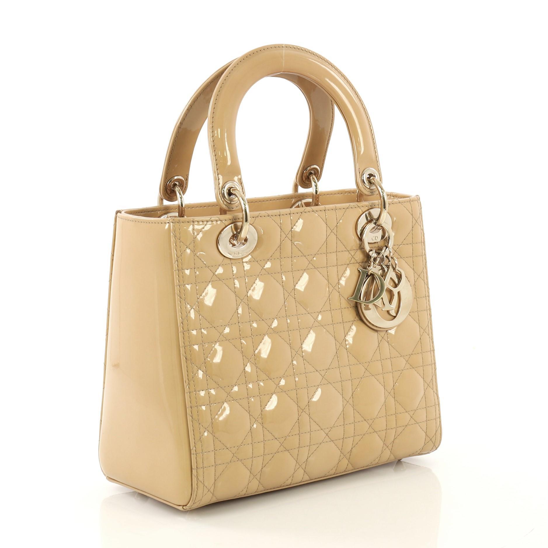 This Christian Dior Lady Dior Handbag Cannage Quilt Patent Medium, crafted in beige cannage quilt patent, features dual top leather handles, Dior charms, and gold-tone hardware. Its zip closure opens to a beige fabric interior with side zip pocket.
