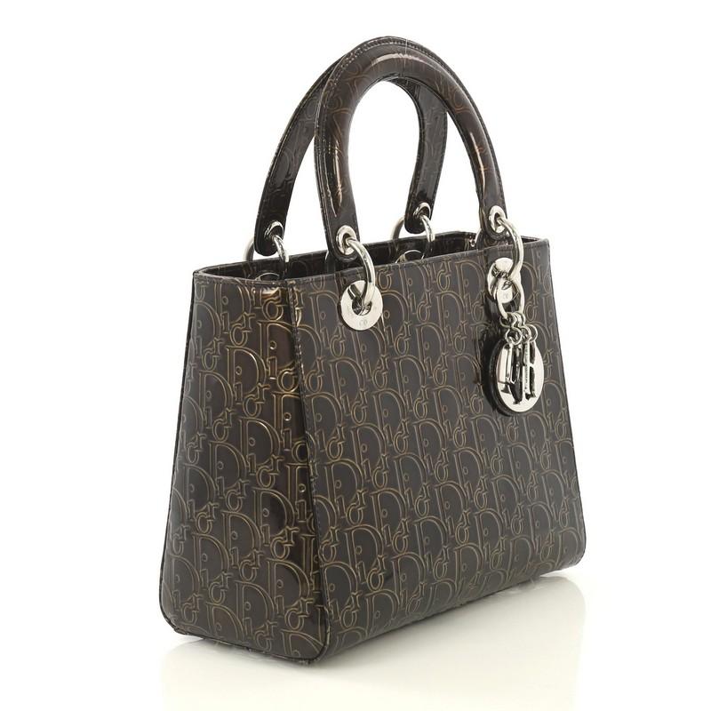 This Christian Dior Lady Dior Handbag Ultimate Embossed Patent Medium, crafted in brown embossed patent leather, features leather top handles, protective base studs and silver-tone hardware. Its zip closure opens to a black nylon interior with zip