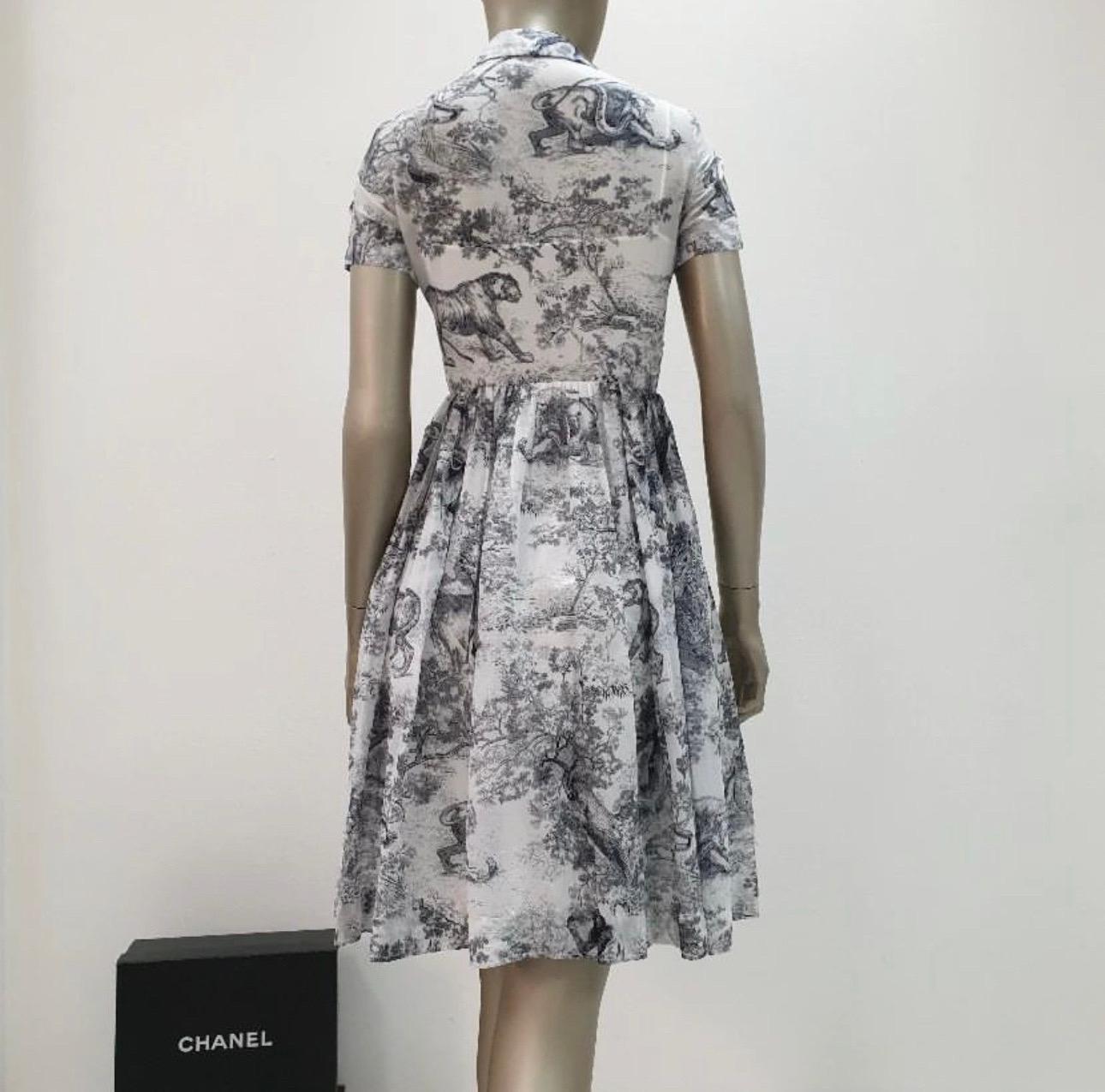 The shirt dress showcases the House's hallmark Toile de Jouy motif in navy blue. Created in cotton voile, it boasts a long and fluid silhouette enhanced by a spread collar and pleated details that highlight the waist. The airy shirt dress can be