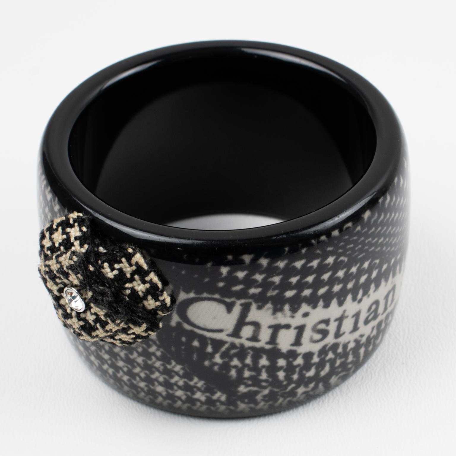 Decorated with a houndstooth print (Pied-de-Poule) and a logo print to the front, this bi-color bangle bracelet from Christian Dior is a bold fashion accessory. This black and white colored resin, acrylic, or Lucite bracelet boasts a lovely woven