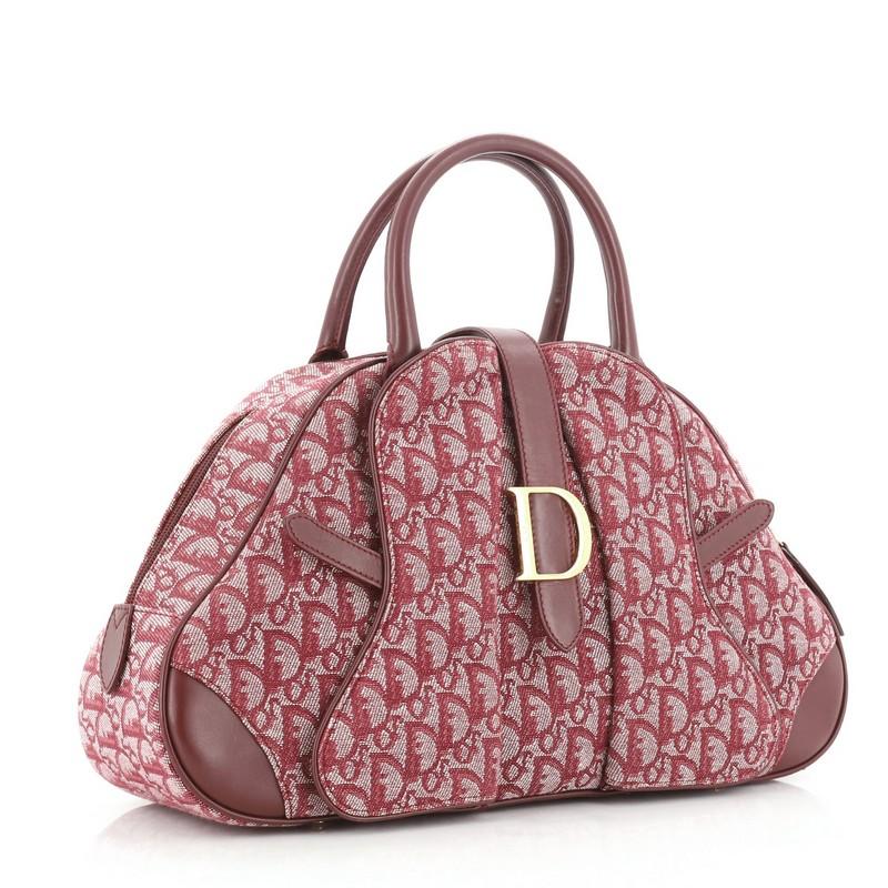 This Christian Dior Saddle Bowler Bag Diorissimo Canvas Medium, crafted from purple diorissimo canvas, features dual rolled leather handles, protective base studs, cross-over strap with Dior clasp, and gold-tone hardware. Its zip closure opens to a