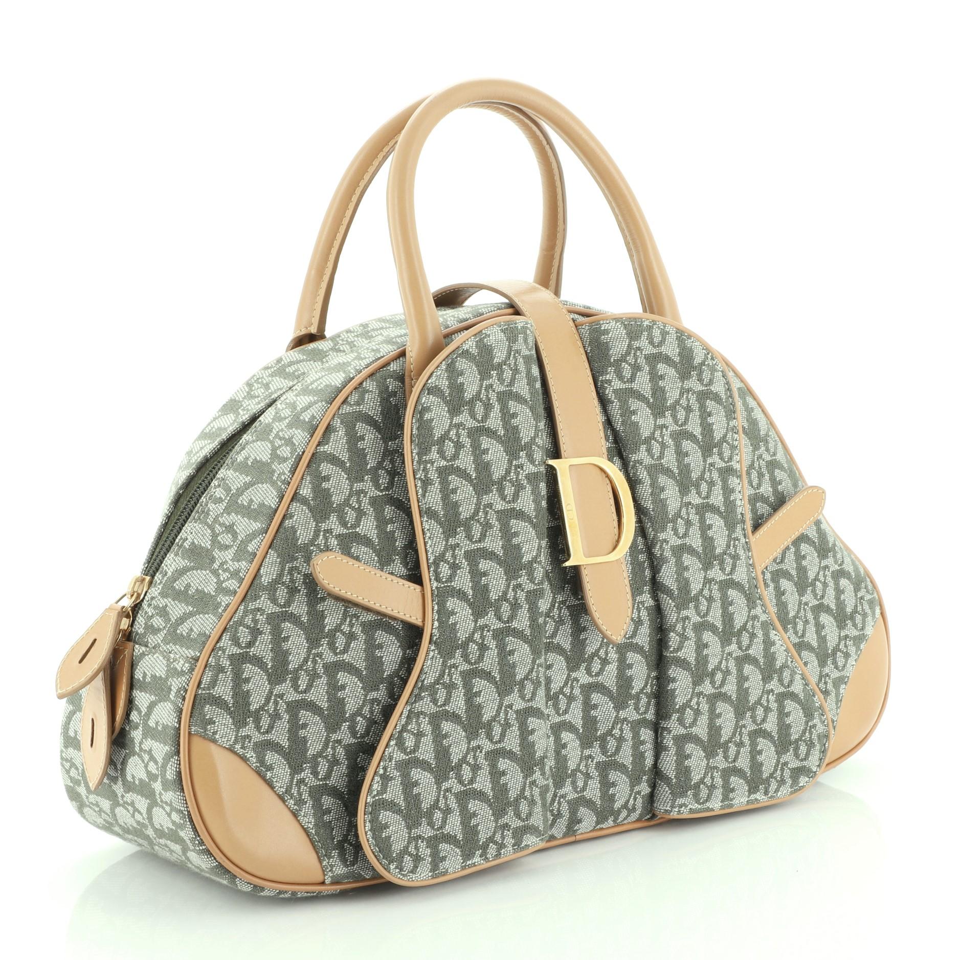 This Christian Dior Saddle Bowler Bag Diorissimo Canvas Medium, crafted from green diorissimo canvas, features dual rolled leather handles, cross-over strap with Dior clasp, and gold-tone hardware. Its zip closure opens to a green nylon interior.