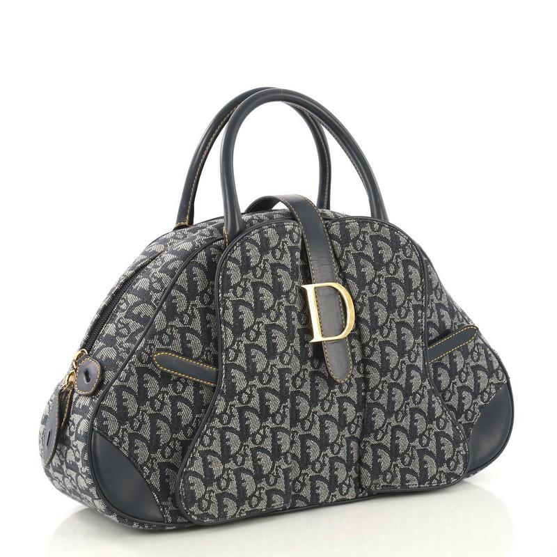 This Christian Dior Saddle Bowler Bag Diorissimo Canvas Medium, crafted from blue diorissimo canvas, features dual rolled leather handles, protective base studs, cross-over strap with Dior clasp, and gold-tone hardware. Its zip closure opens to a