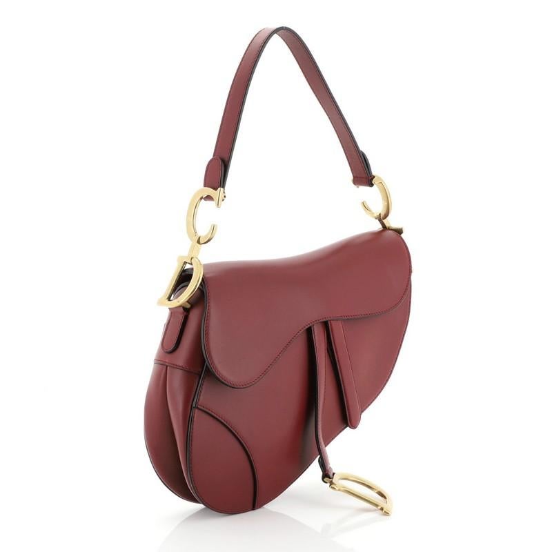 This Christian Dior Saddle Handbag Leather Medium, crafted from red leather, features a top handle adorned with metal 'CD' hardware and aged gold-tone hardware. Its fold over top opens to a red suede interior with zip pocket.

Estimated Retail