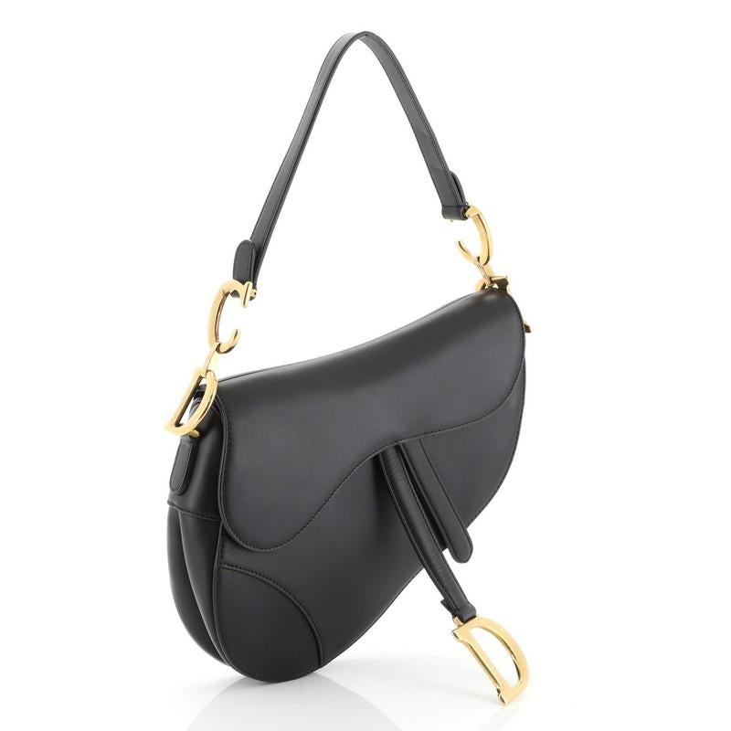 This Christian Dior Saddle Handbag Leather Medium, crafted from black leather, features a top handle adorned with metal 'CD' hardware and aged gold-tone hardware. Its fold over top opens to a black suede interior with zip pocket. 

Condition: Great.