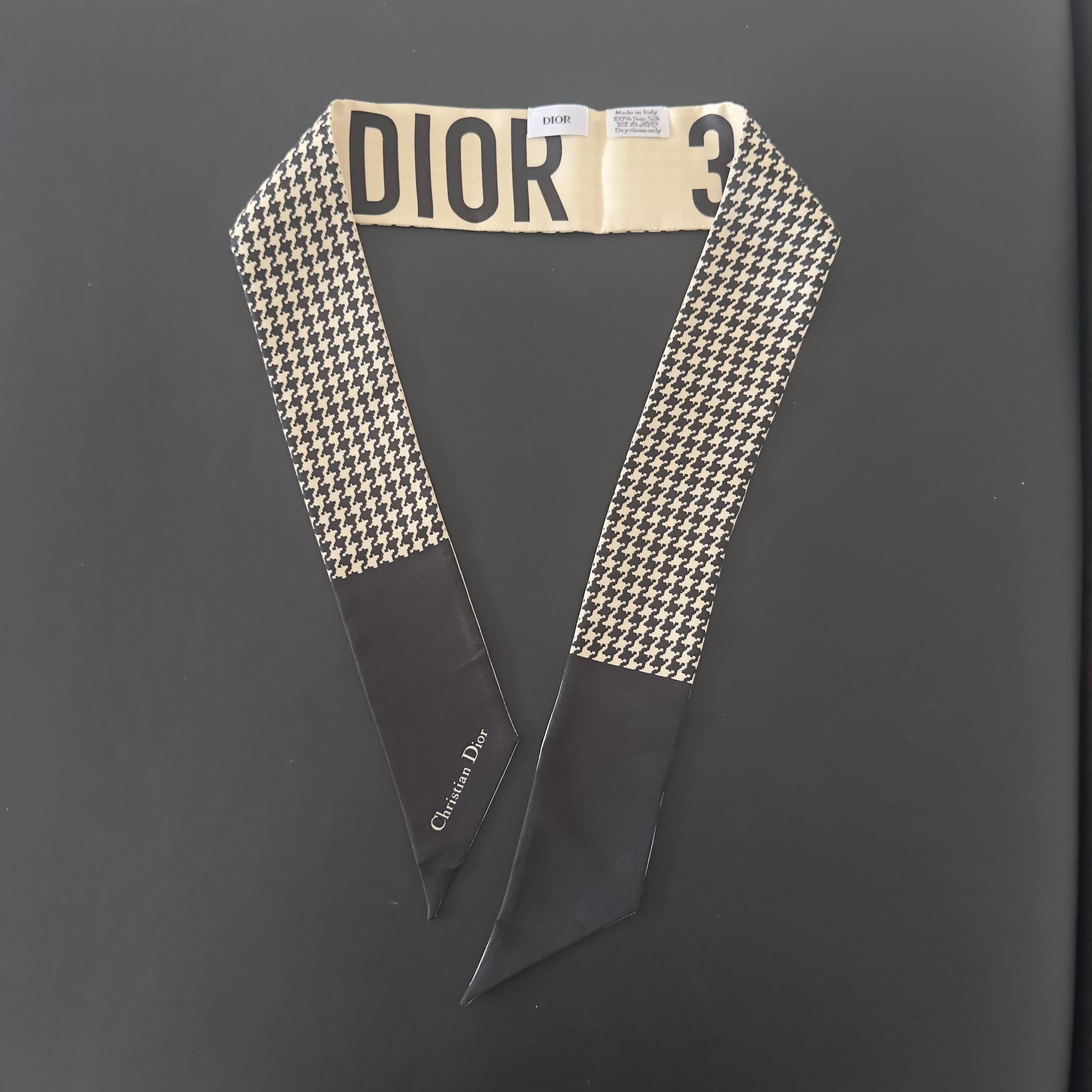 Une icône de la Maison réinterprétée dans une tonalité contemporaine.
Ce twilly Dior est un accessoire de luxe extrêmement polyvalent, réalisé en 100 % soie avec une double impression : d'un côté le célèbre motif pied-de-poule noir et beige, de