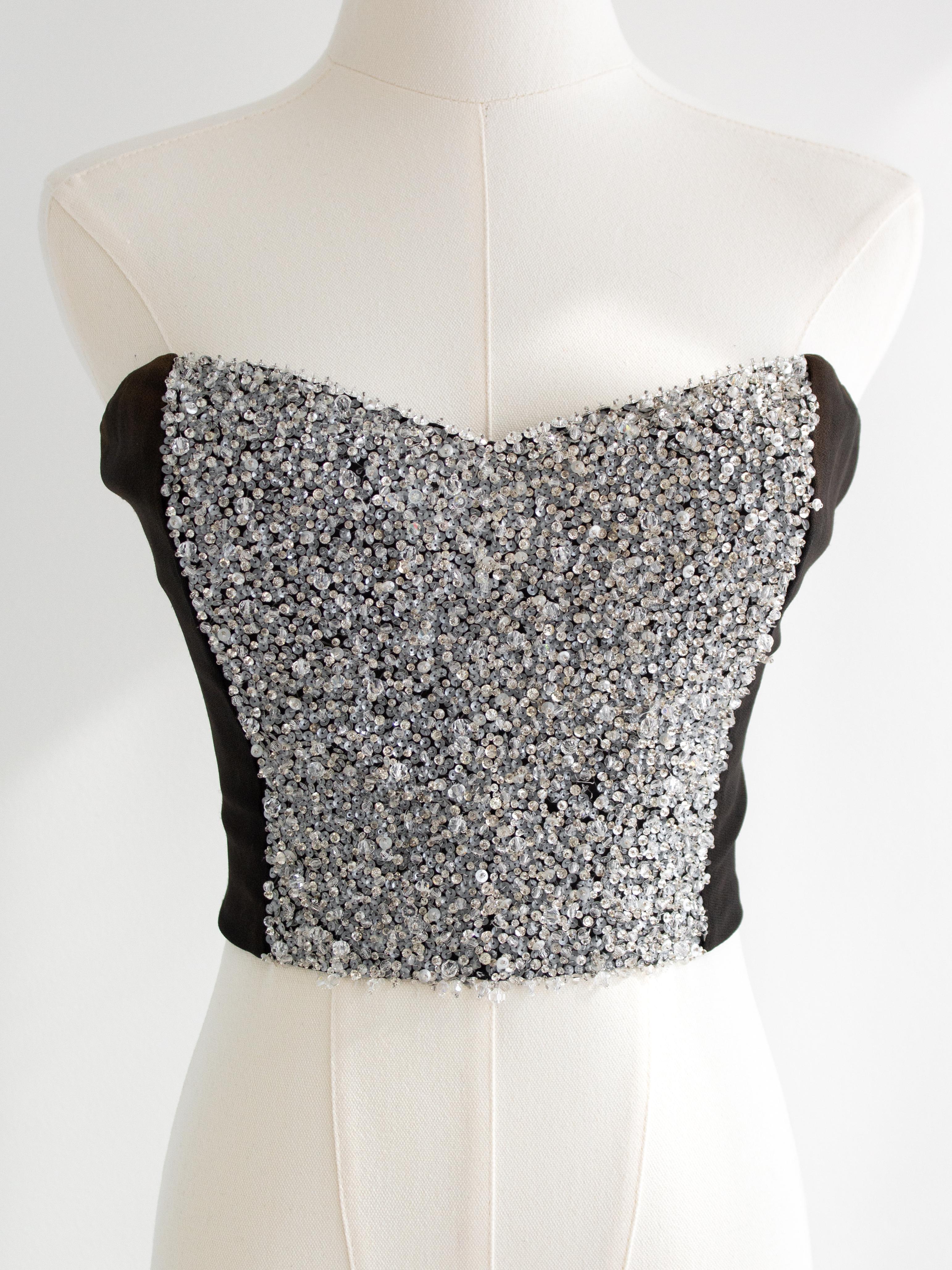 This stunning vintage bustier from Christian Dior’s S/S 1997 collection features an elegant black body adorned with intricate silver embellishments. The sweetheart neckline is accentuated by a shimmering array of crystals and sequins, adding a