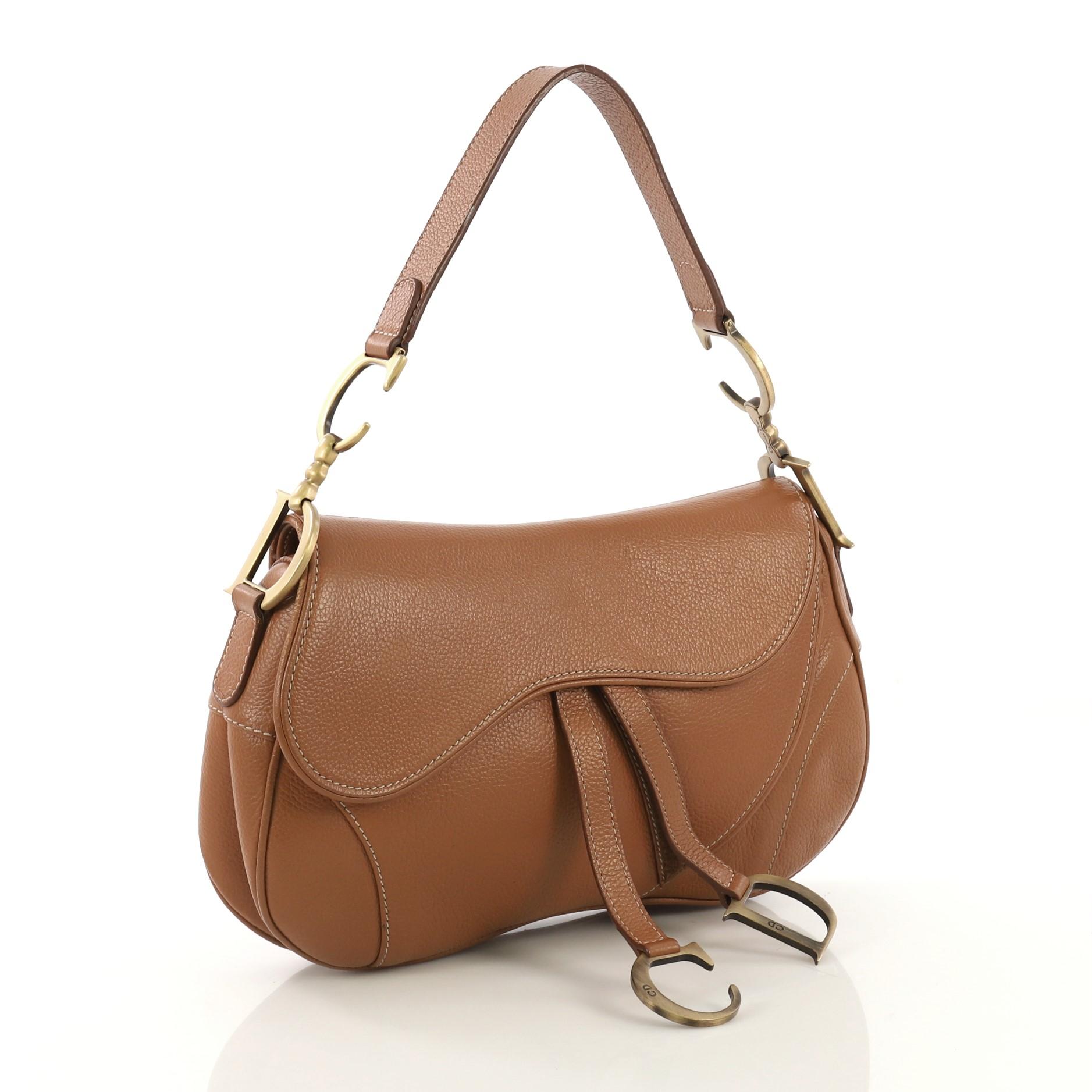This Christian Dior Vintage Double Saddle Bag Leather, crafted from brown leather, features a top handle adorned with metal 'CD' finishing and aged gold-tone hardware. Its fold over top opens to a dark brown fabric interior with zip pocket.