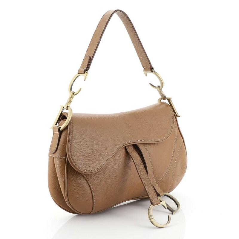 This Christian Dior Vintage Double Saddle Bag Leather, crafted from brown leather, features a top handle adorned with metal 'CD' finishing and aged gold-tone hardware. Its fold over top opens to a brown fabric interior with zip pocket. 

Condition: