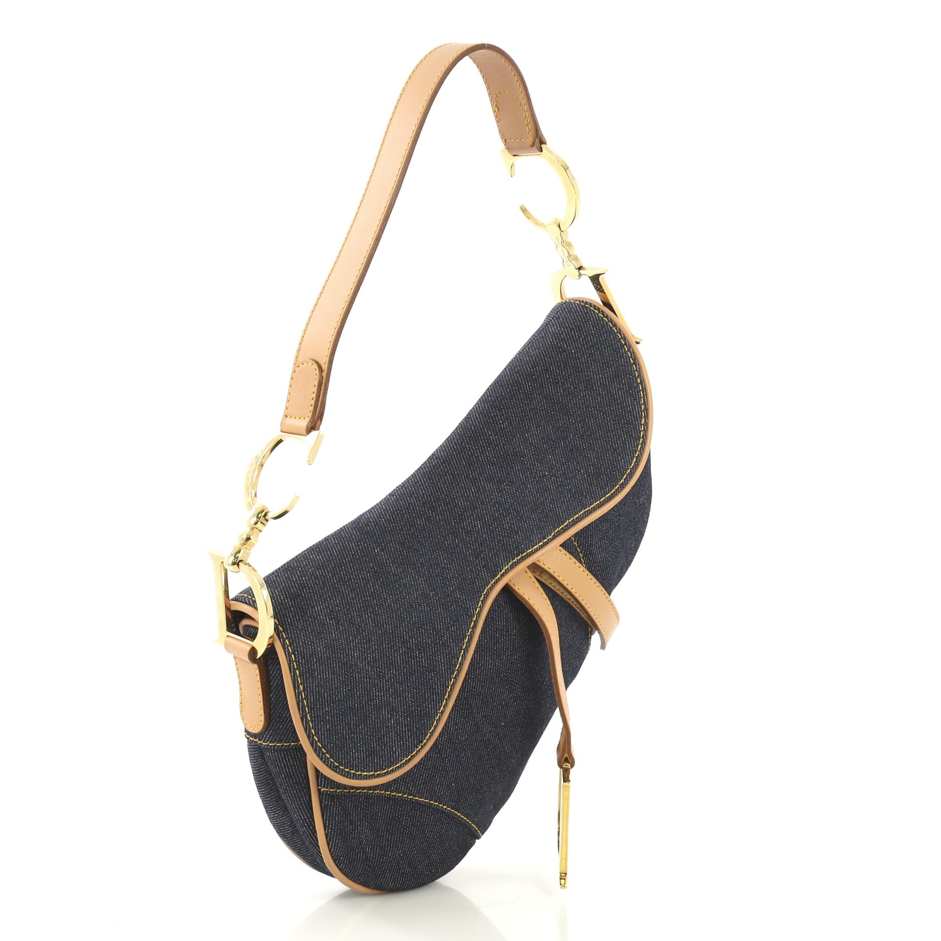 This Christian Dior Vintage Saddle Bag Denim Medium, crafted from blue denim, features a top handle adorned with metal 'CD' finishing, saddle-shaped silhouette, and gold-tone hardware. Its fold over top opens to a white printed fabric interior with