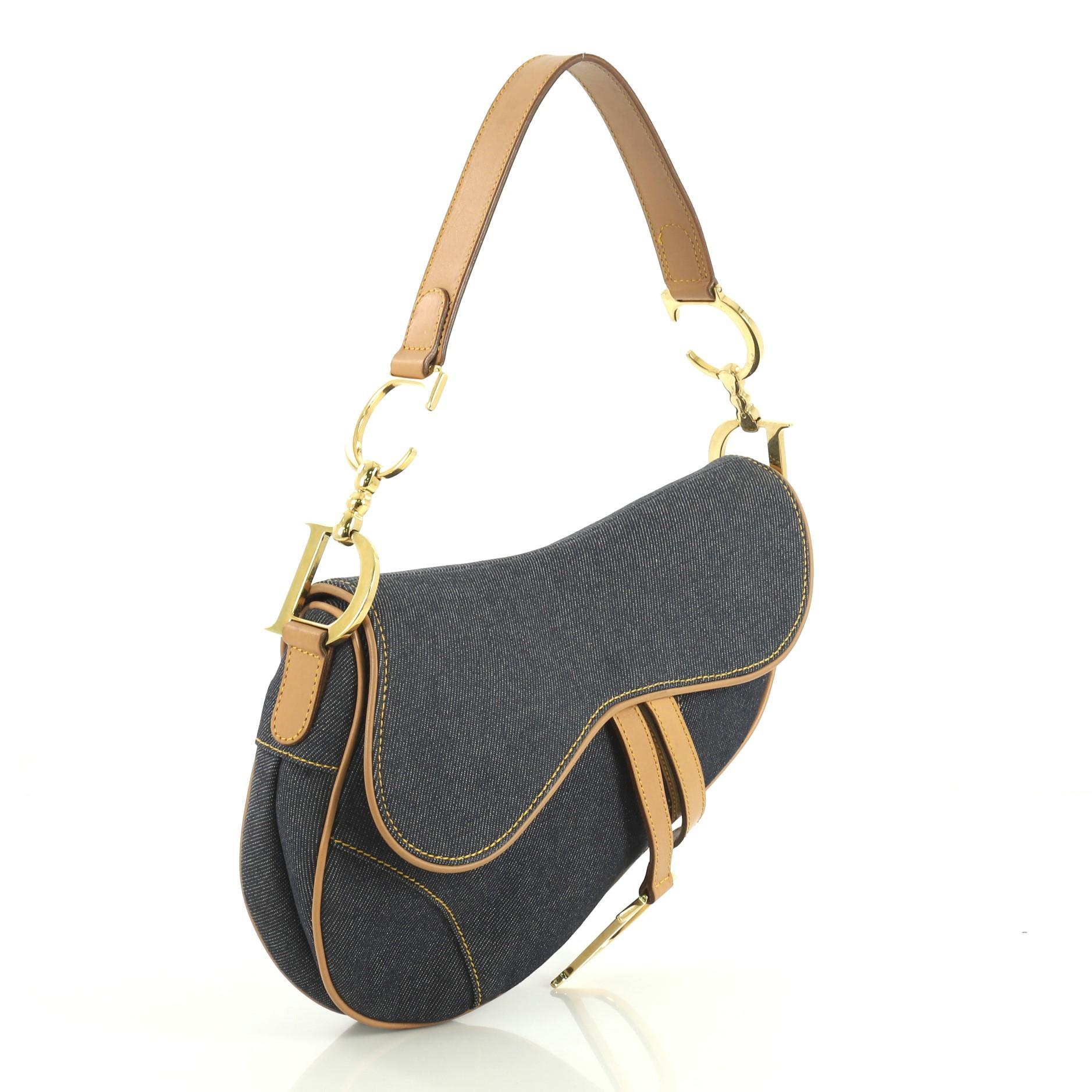 This Christian Dior Vintage Saddle Bag Denim Medium, crafted from blue denim, features a top handle adorned with metal 'CD' finishing, saddle-shaped silhouette, and gold-tone hardware. Its fold over top opens to a blue nylon interior with zip