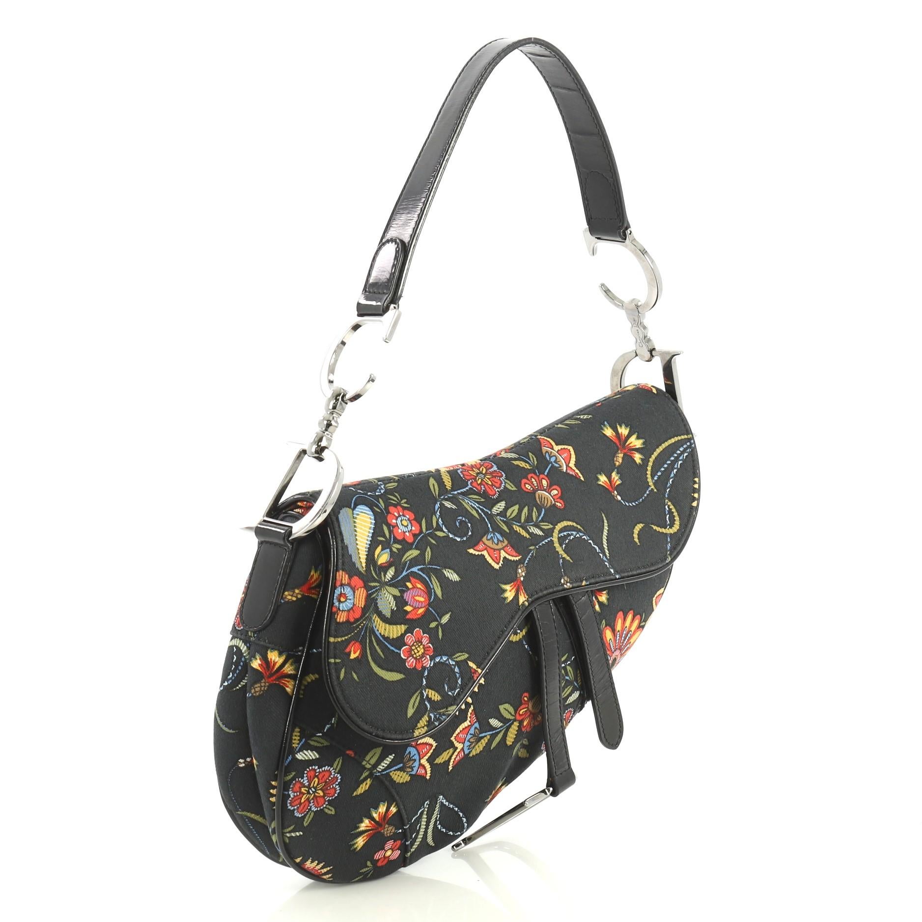 This Christian Dior Vintage Saddle Bag Printed Canvas Medium, crafted from black multicolor printed canvas, features a top handle adorned with metal 'CD' finishing, floral print, saddle-shaped silhouette, and silver-tone hardware. Its fold over top