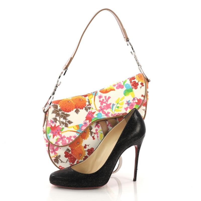 This Christian Dior Vintage Saddle Bag Printed Leather Medium, crafted from white printed leather, features a top handle adorned with metal 'CD' finishing, bright floral print, saddle-shaped silhouette, and silver-tone hardware. Its fold over top