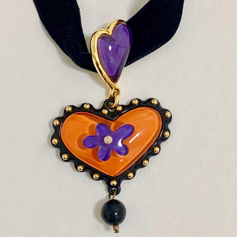 Christian Lacroix resin heart pendant.  We can find in the center a daisy resin purple color and a small pearl. This heart is attached to a purple resin heart. The link is a black velvet ribbon.
It comes from the Summer 1994 collection. Made in