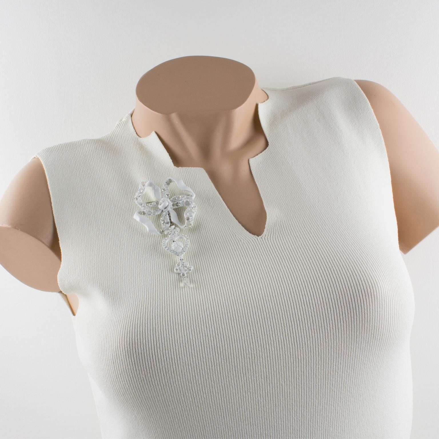 Cette broche signée Christian Lacroix Paris, rare et étonnante, présente un nœud pendentif surdimensionné avec un cœur ex-voto, une tête de mort et des breloques du logo CL en métal émaillé blanc orné de strass en cristal clair et en cristal