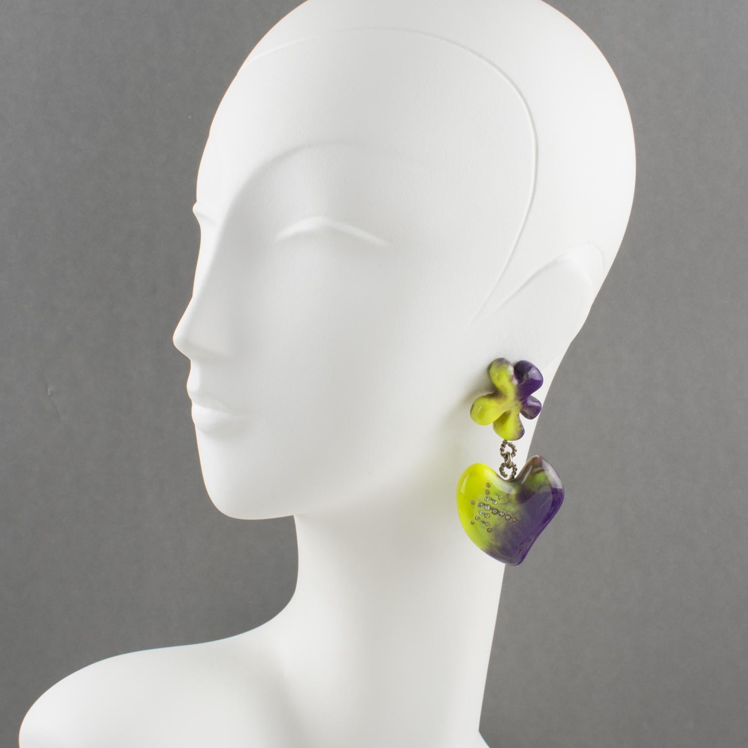 These stunning Christian Lacroix Paris clip-on earrings feature a massive resin dangling shape with abstract flowers and a heart design ornated with clear crystal rhinestones. The pieces boast a fabulous palette of amethyst purple with pistachio