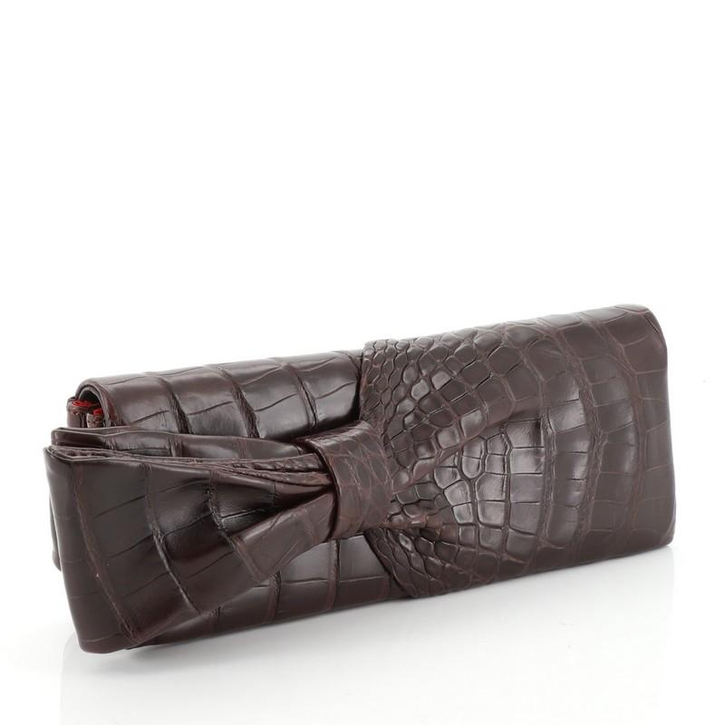 This Christian Louboutin Aionoeud Bow Clutch Crocodile, crafted in genuine brown crocodile skin, features a prominent bow on the flap. Its magnetic snap button closure opens to a red fabric interior. . This item can only be shipped within the United