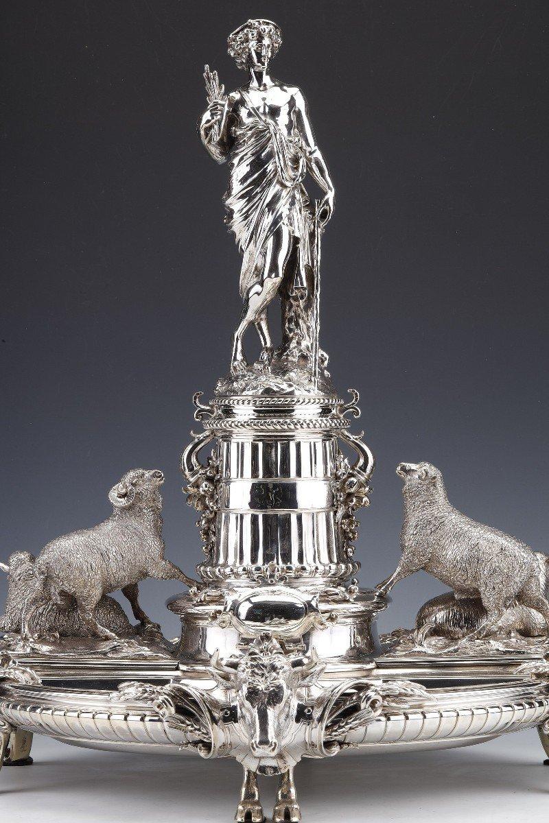 Solid silver centerpiece in a round shape, placed on rolling clog feet. The subject is a decoration of sheep as well as heads of bulls framing a sculpture of a Shepherd dressed in the antique, probably CERES symbolizing earthly fertility. This