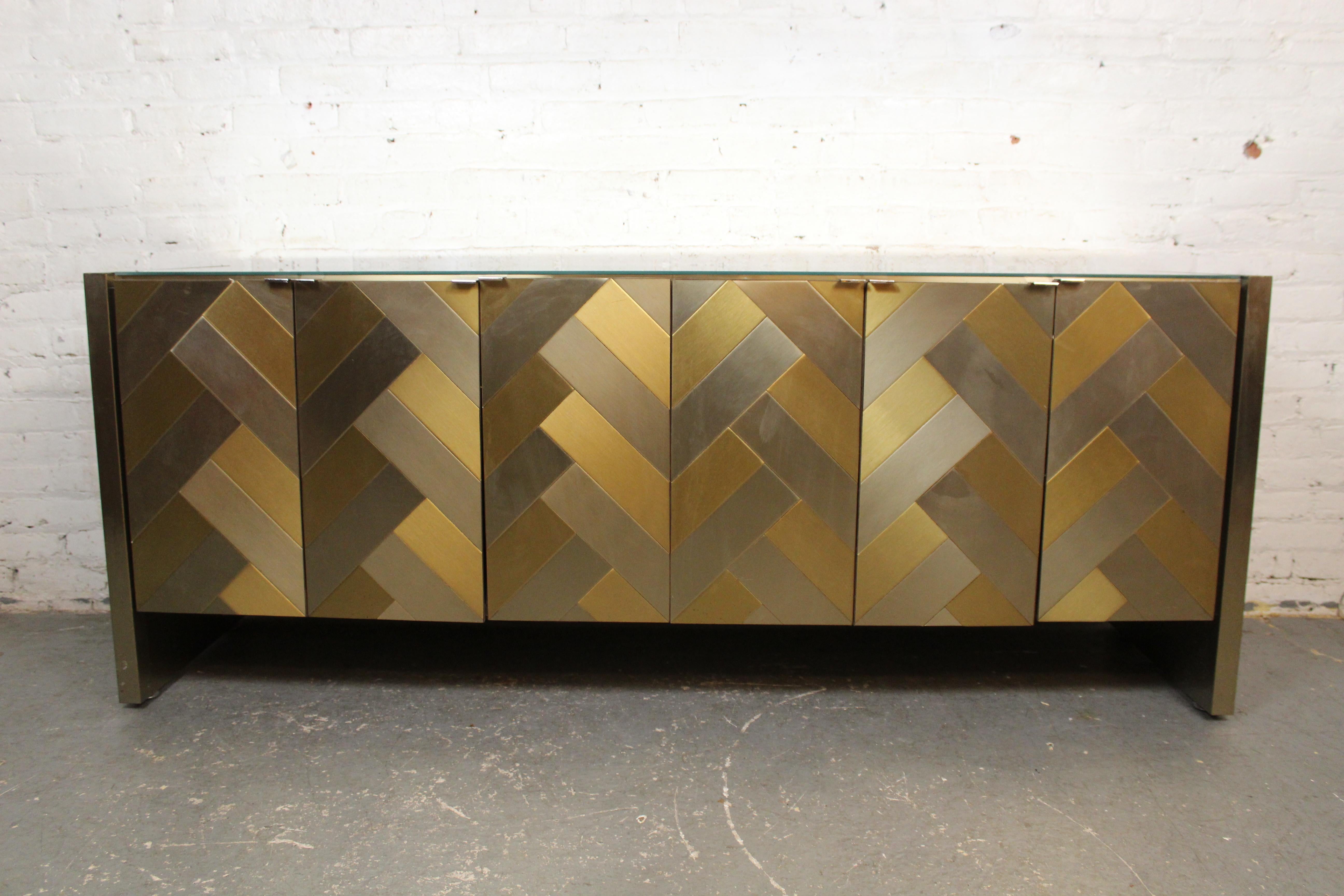 This stunning cabinet by Rockford, Illinois' Ello Furniture features an inlaid patchwork design made of brushed brass and chrome tiles complimenting an oversized mirrored top. Large accordion doors fold open to reveal two spacious cabinets with