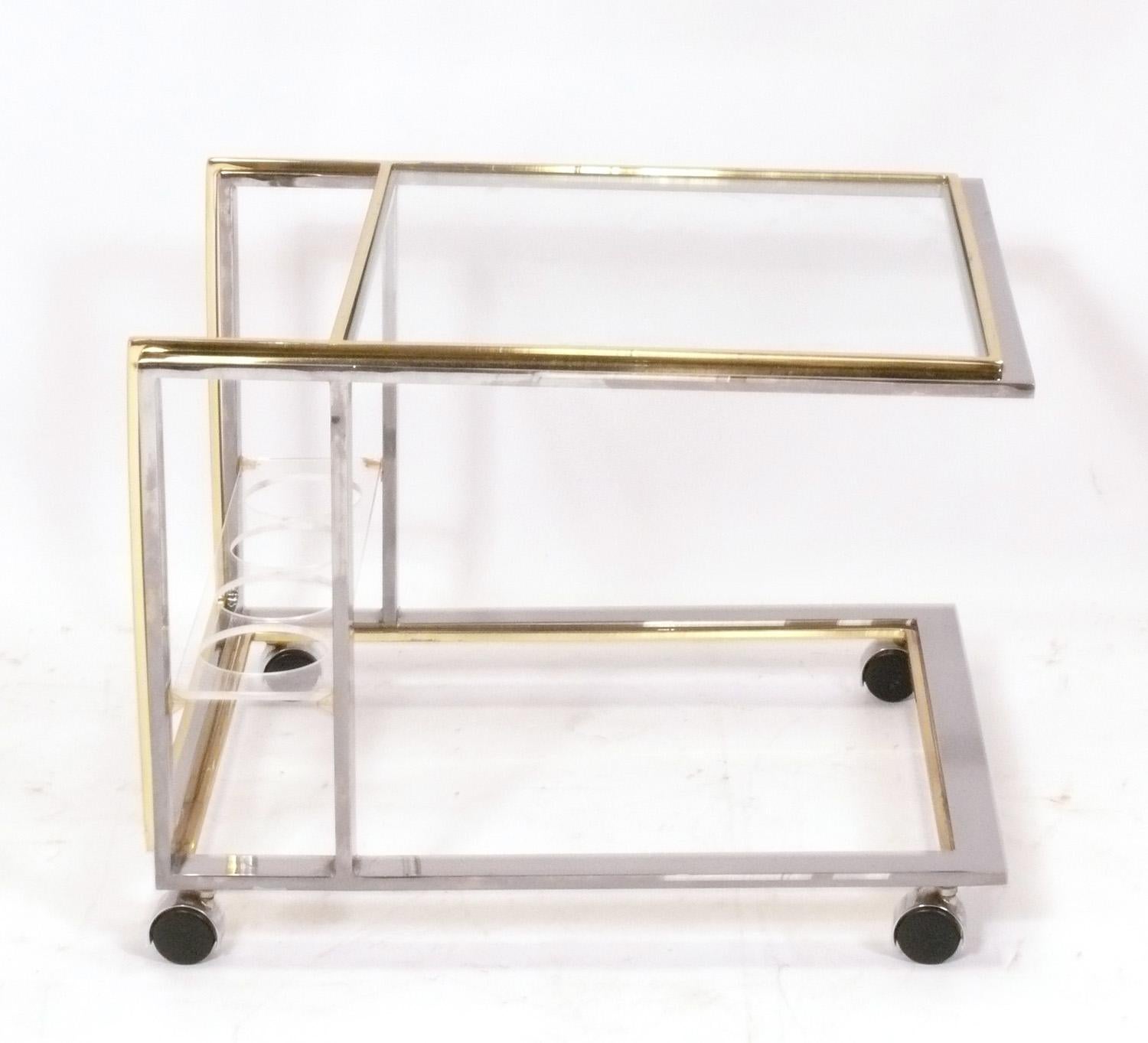 Chrome and Brass Mid Century Italian Bar Cart, attributed to Romeo Rega, Italy, circa 1960s. It has two glass shelves and a lucite bottle rack at the bottom. The lower glass panel was broken while preparing this cart to photograph and will be