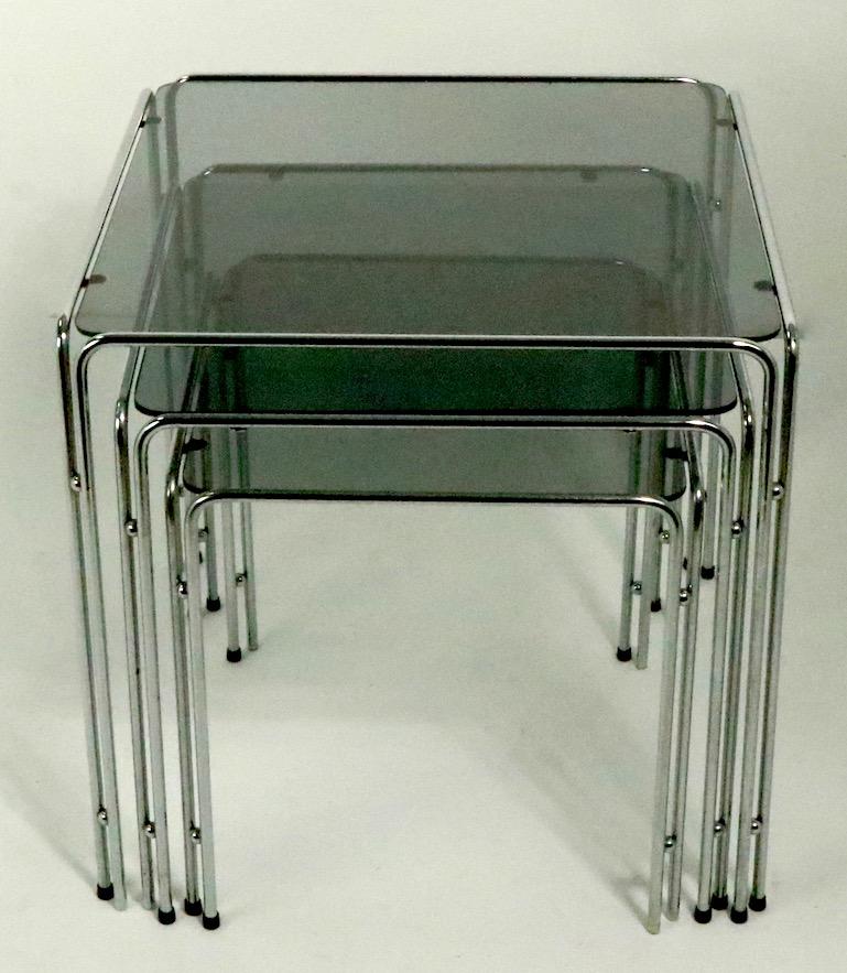 Chic set of three chrome and glass nesting tables circa 1970s. Each table has a bright chrome base with parallel chrome rod legs with decorative chrome ball accent joinery, the tops are original gray glass with rounded corners. Al are in very good