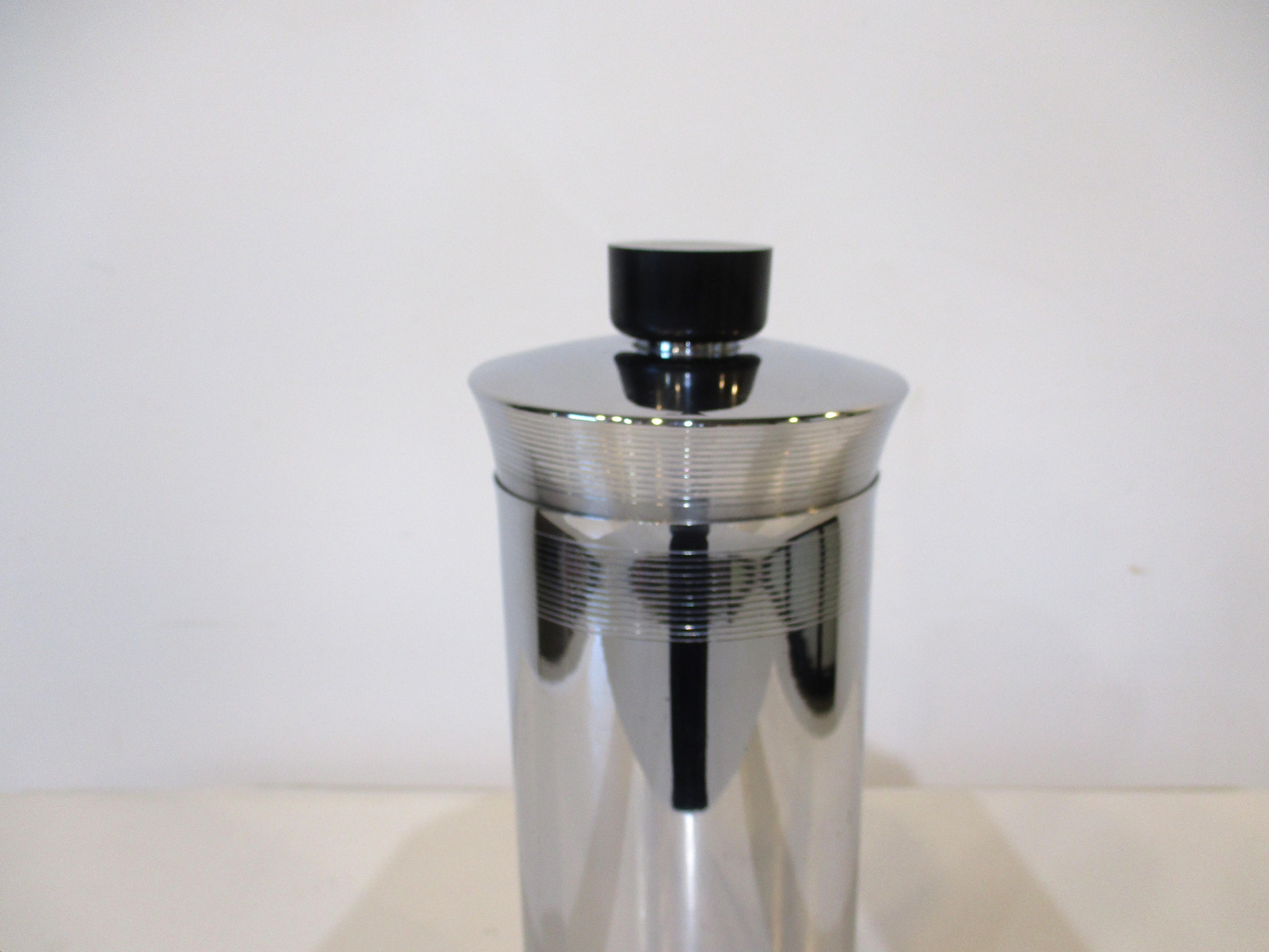 A chrome Art Deco cocktail shaker with ribbed grooves to the top and bottom section of the container. The lid has matching grooves with built in strainer topped with a black bakealite cap. Manufactured and designed in the manner of Manning Bowman.