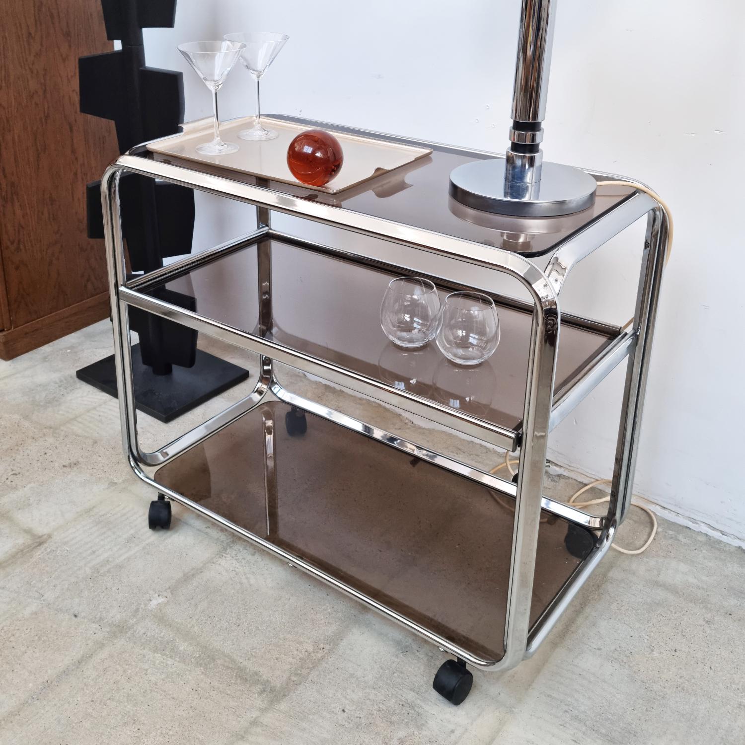 Introducing our Chrome Bar Cart from the 1970s, a true embodiment of vintage charm. Crafted with a sleek chrome frame and complemented by smoked glass shelves, this piece exudes an air of sophistication. The middle shelf offers versatility with its