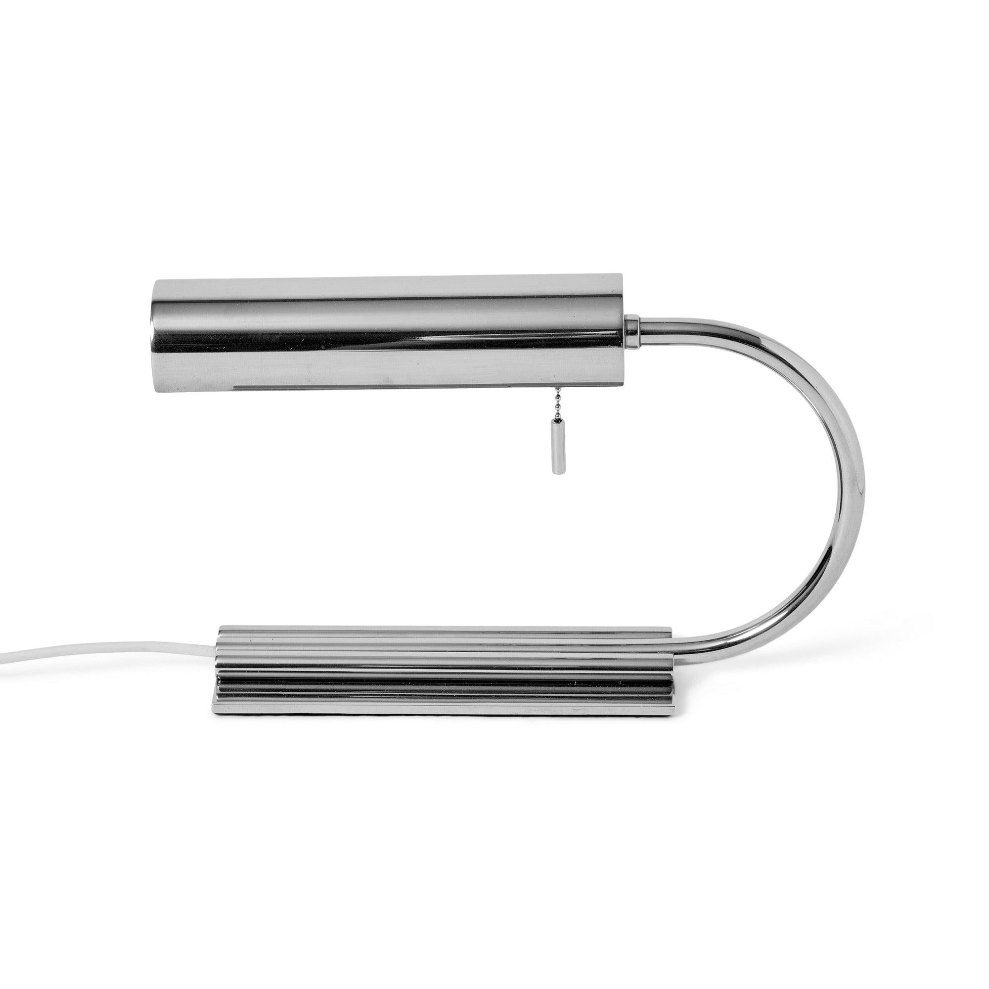 A low chrome desk lamp consisting of three chrome tubes set on a flat, rectangular base the center tube arcing up over the base suspending a cantilevered cylindrical shade. No identifying markings.