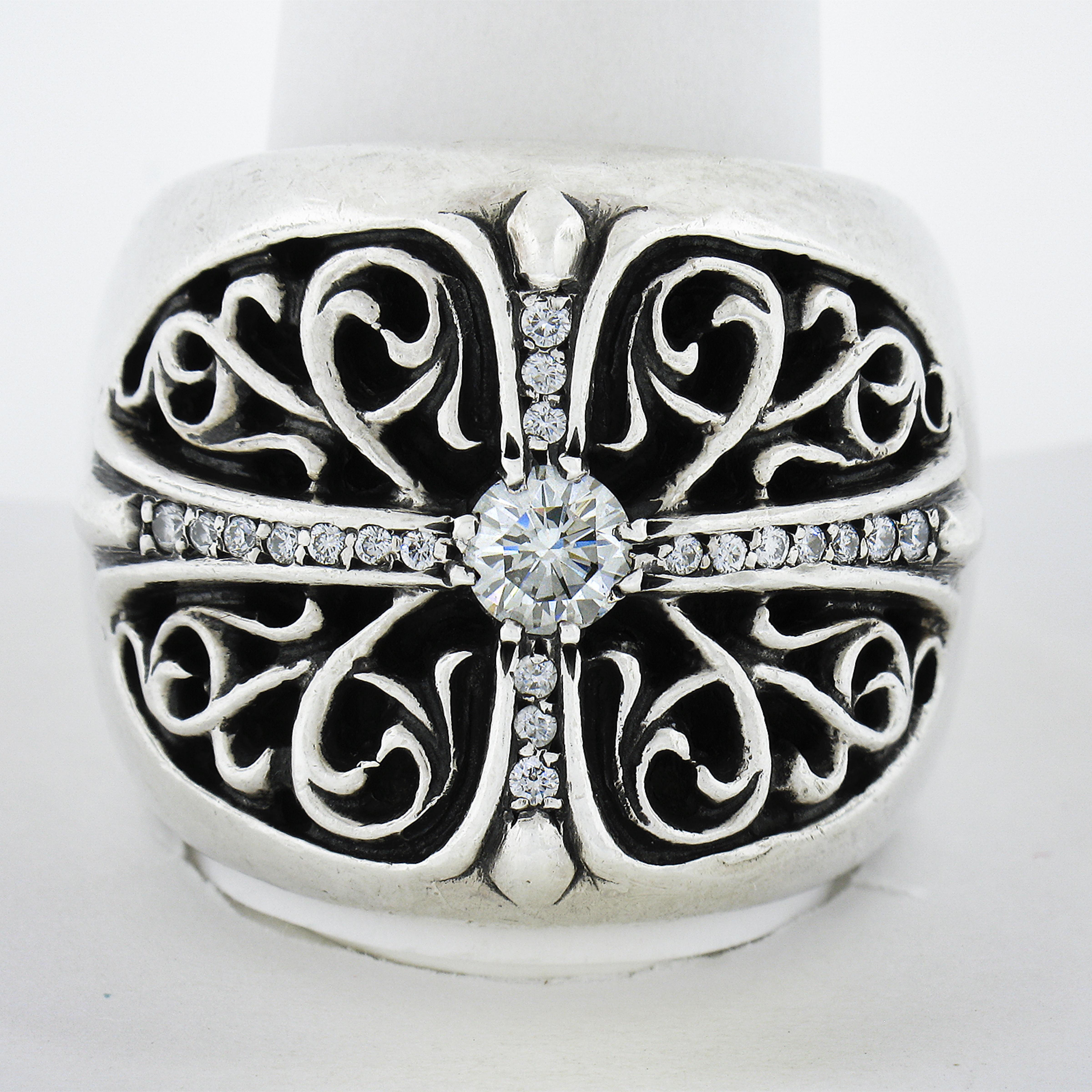 This bold and sharp-looking men's ring by Chrome Hearts is solidly crafted in sterling silver and and features a wide design with detailed scroll open work and is adorned with 0.77 carats of truly fine quality diamonds throughout. The center diamond