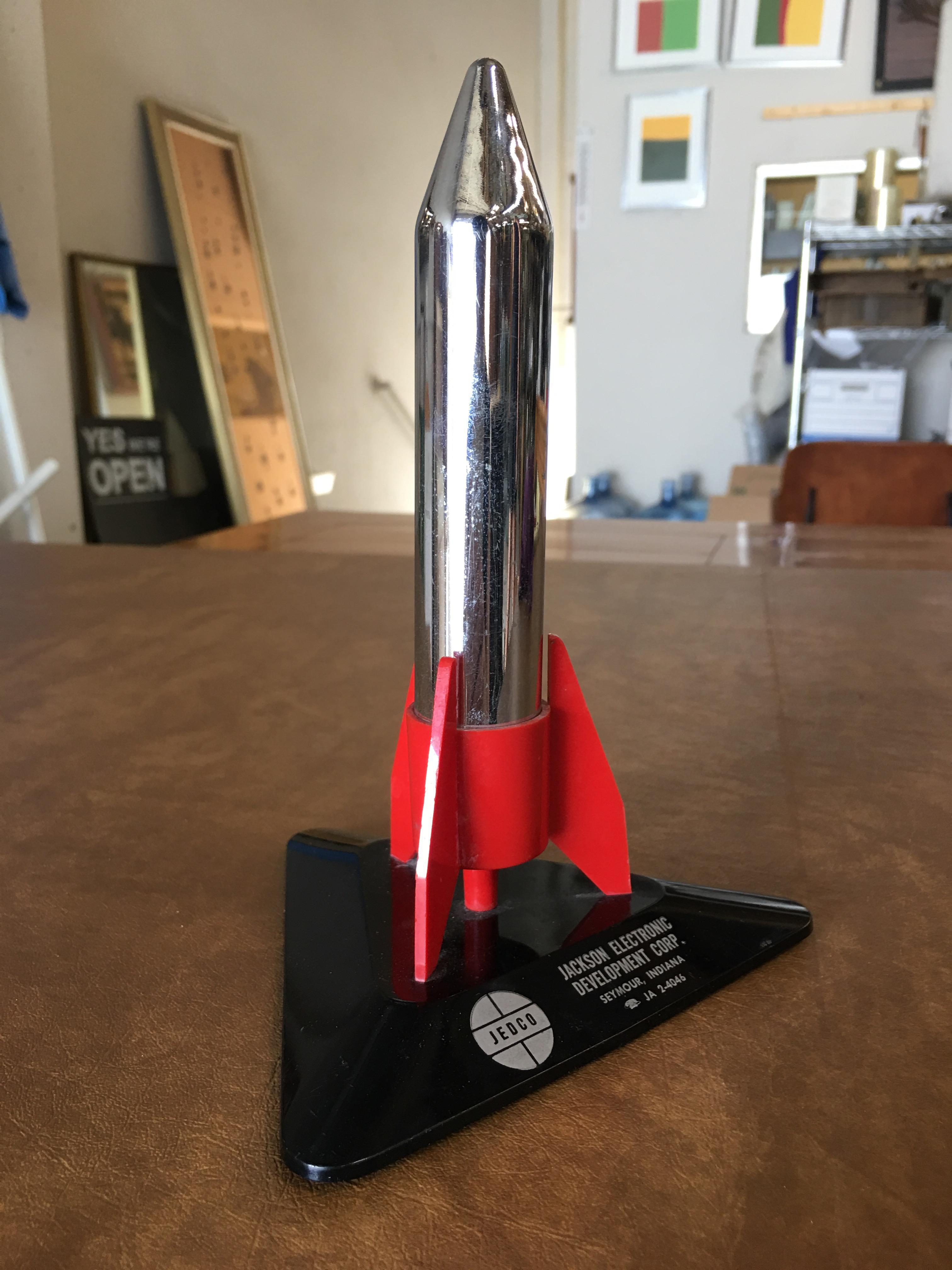 Vintage 1960 chrome Jedco rocket shaped tabletop lighter featuring a chrome rocket ship resting in a black and red plastic base. The front text reads 