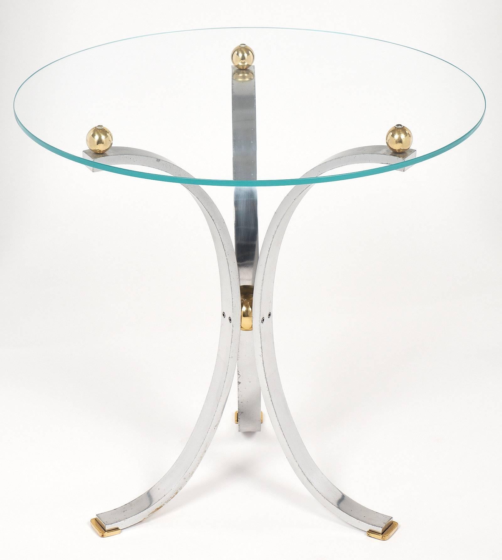 Table d'appoint chromée moderniste française avec une base tripode en forme de guéridon formée par trois arcs en acier chromé. La base est terminée par des pieds en laiton à leur base, et maintiennent le plateau en verre avec trois sphères en laiton