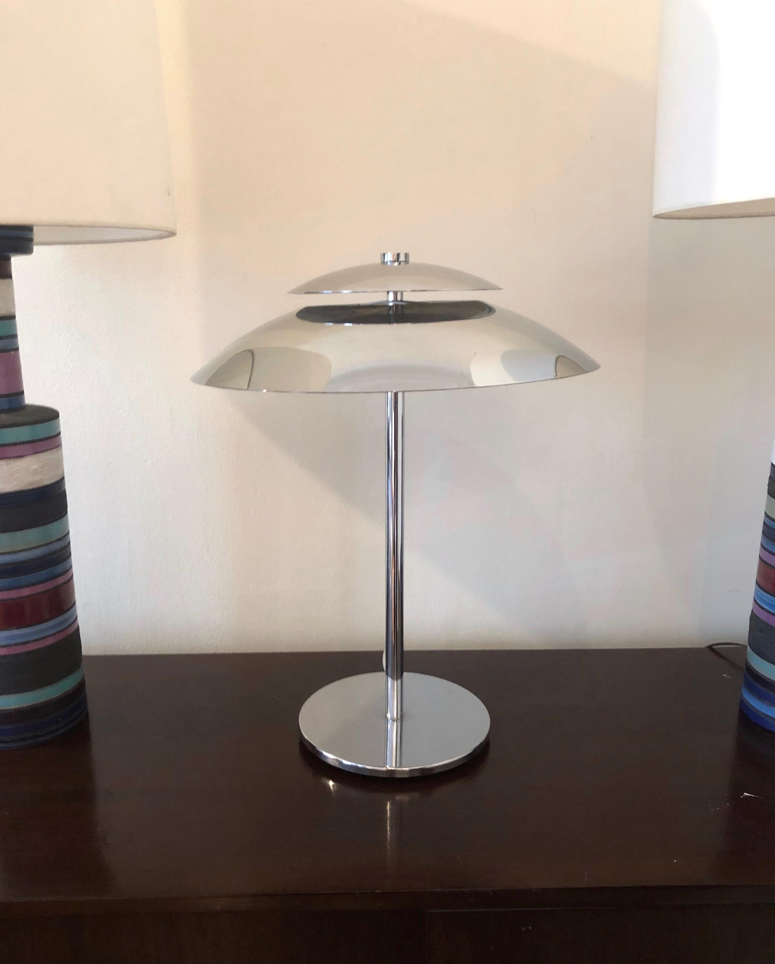 A well made, vintage modern all-chrome table lamp has a round, weighted base and cylindrical stem that hold the rounded triangular shade featuring an elevated segment. The lamp has two sockets, a flat round finial, and is made of sturdy gauge metal