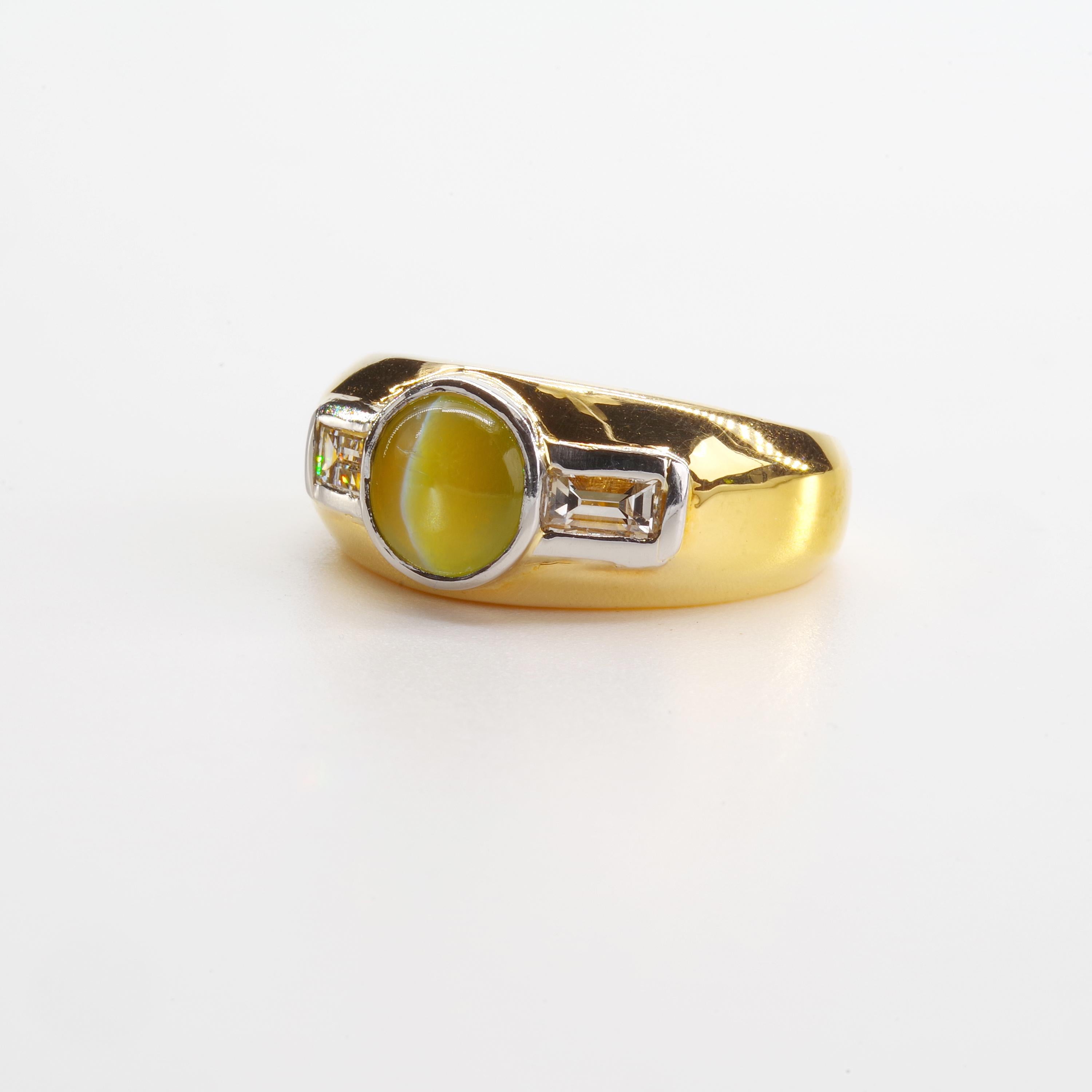 This sleek, handsome 14k yellow gold mid-century ring features a glowing, honey-colored chrysoberyl cat's-eye of approximately 2 carats, bezel-set in 14k white gold and flanked on either side—also set into white gold—by a pair of clean, bright-white