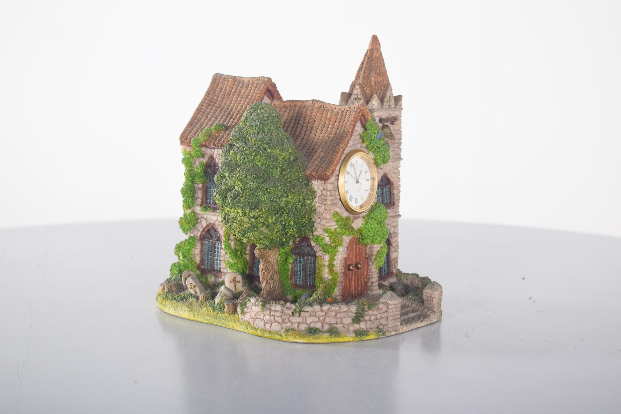 Decorative clock in the shape of a church. It represents a picturesque miniature church, richly detailed to evoke traditional European architecture. Made of resin, it features a tower with a small bell, arched windows, wooden doors and a