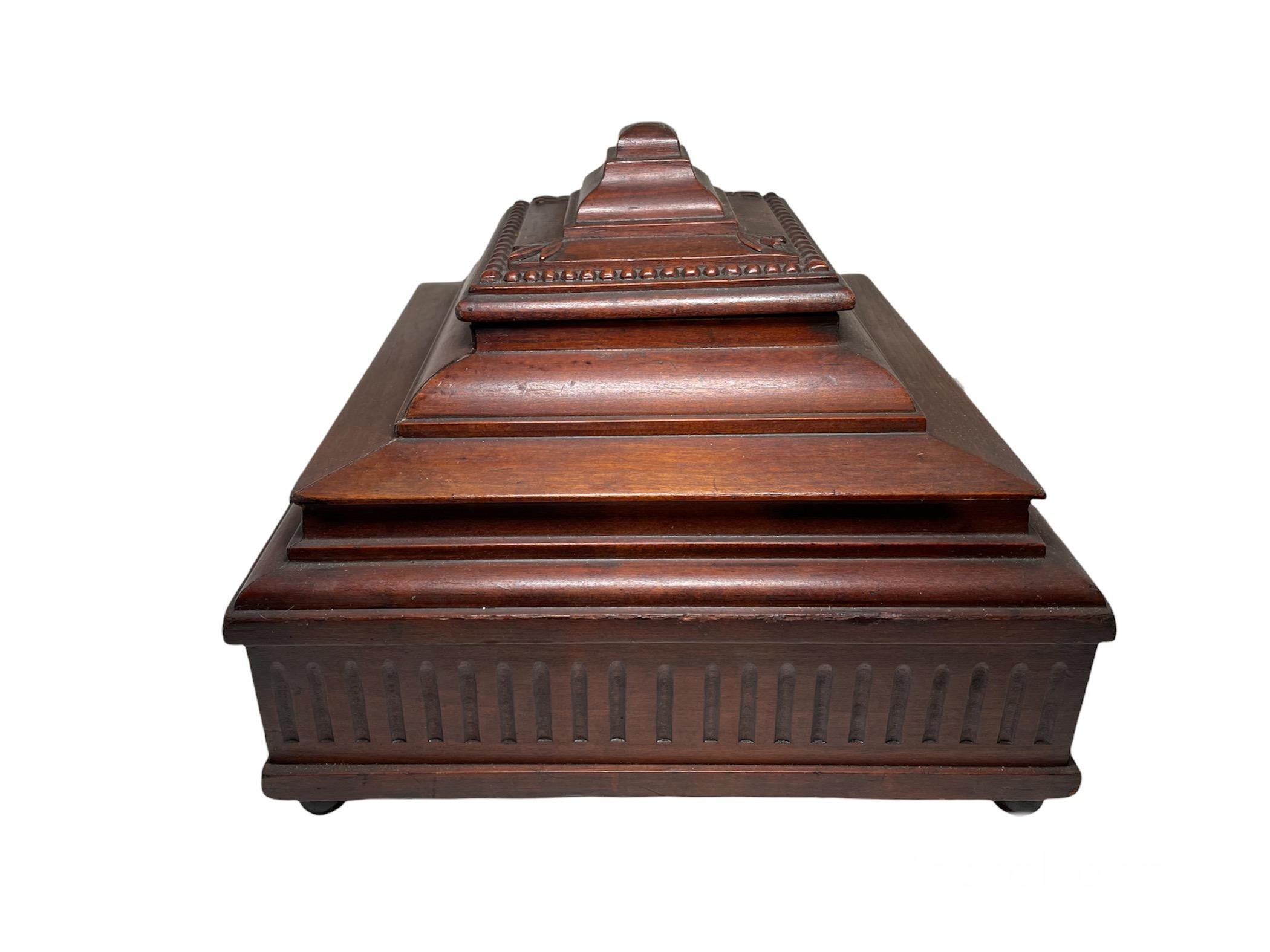 This is a cigar humidor wooden box shaped as a pagoda. The top of the lid is adorned with some relief of leaves in the corners and some beaded chain in its border. Also the wooden square bottom border has some fluted designs. The inside case has a