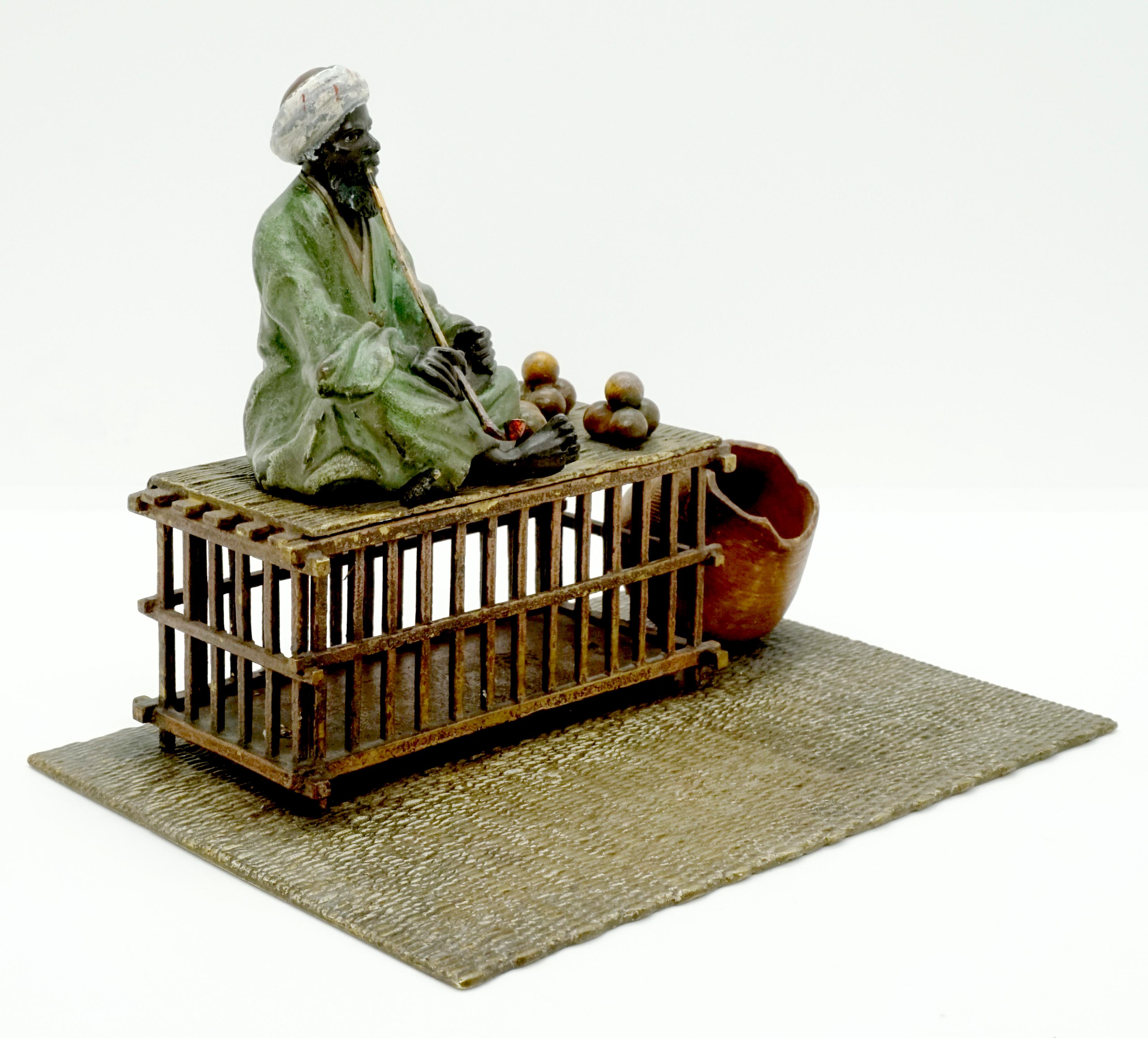 Excellent piece of Viennese bronze Art, circa 1900:
A bearded oriental in a long green robe and turban sits cross-legged on a hinged wooden cage on a carpet and smokes a long pipe. Next to him are piled balls that could be oranges, next to the cage