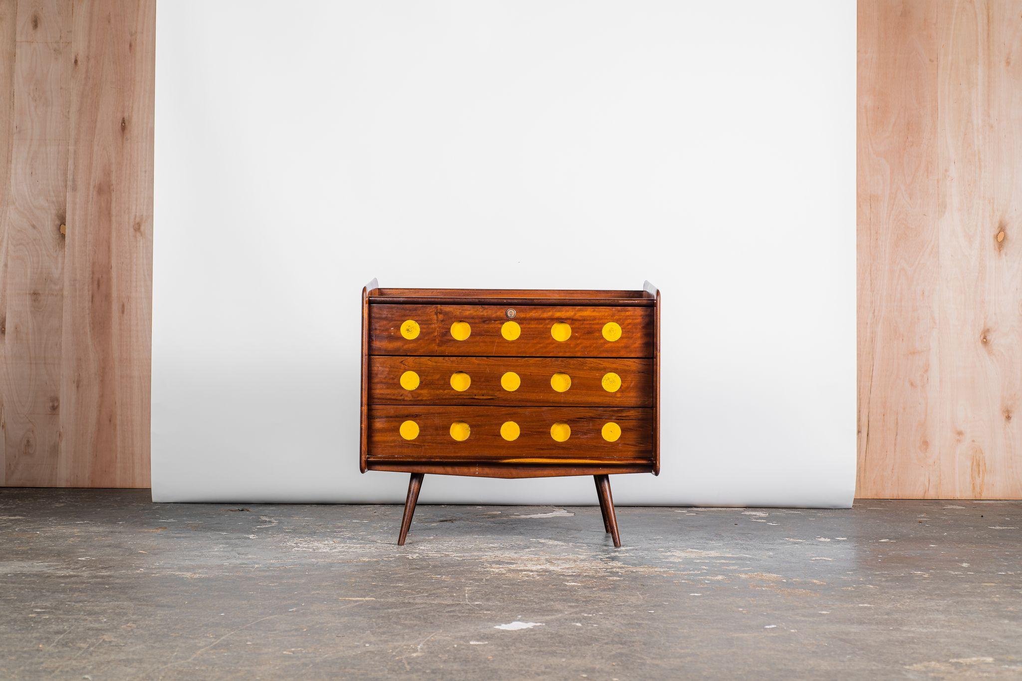 This three-drawer dresser crafted from solid hardwood adorned with vibrant yellow circle motifs exemplifies the whimsical furniture creations by Móveis Cimo during the 50s. Originally conceived as part of a youth's room collection, this dresser