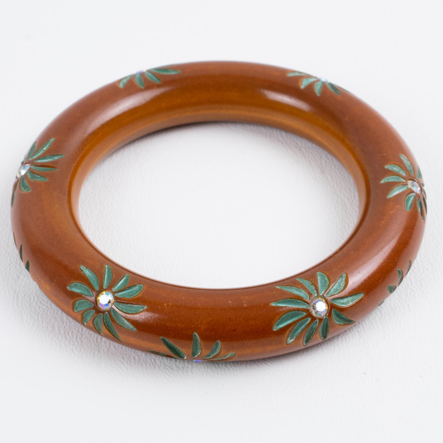 This stunning cinnamon-brown Bakelite bracelet bangle has a chunky, rounded domed shape and floral carving around it. The carved floral design is enhanced with contrasting green paint and AB crystal rhinestones. The piece boasts an intense